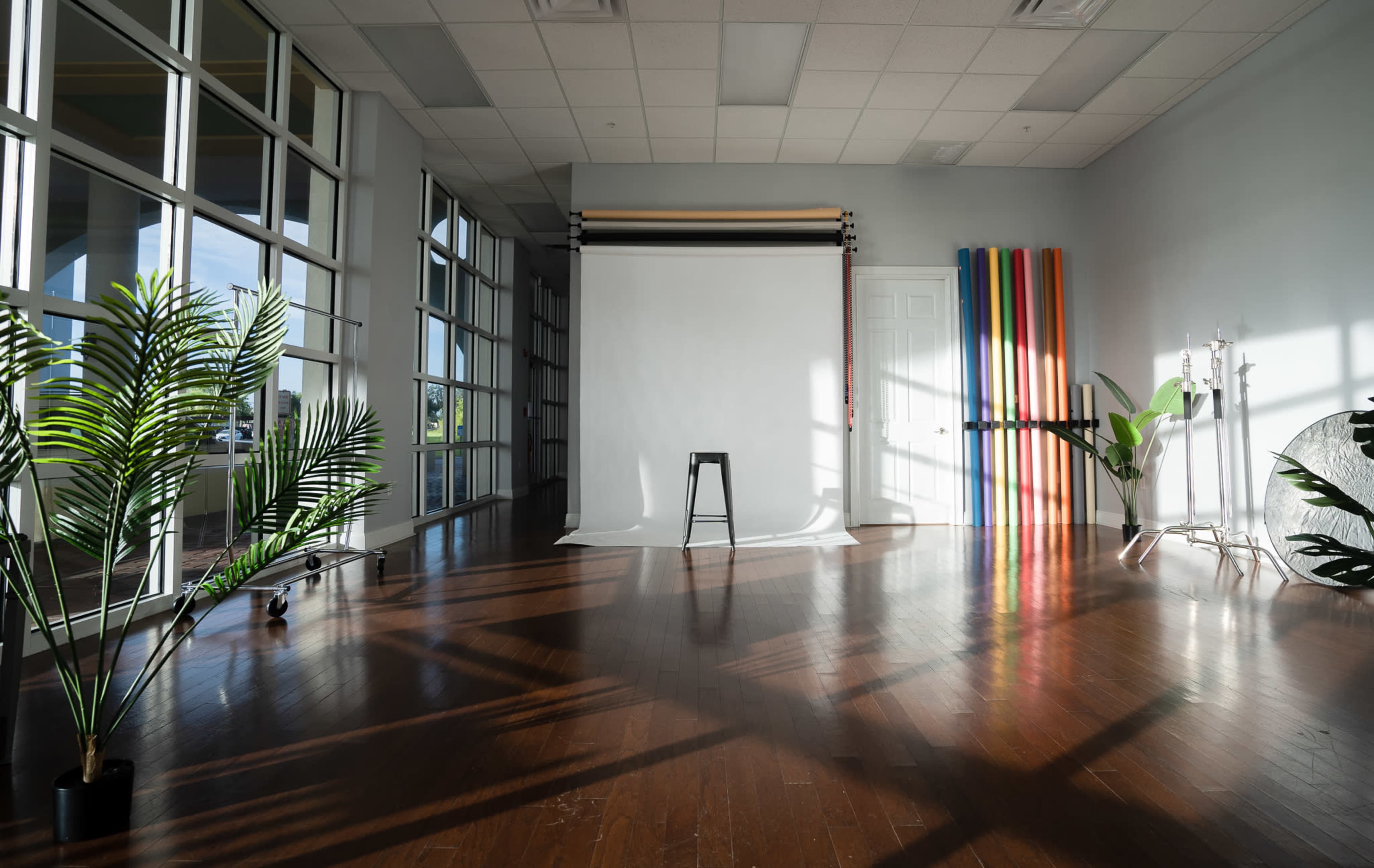 The image shows a spacious studio with a white backdrop, a black stool in the center, and colorful vertical stripes against a wall, illuminated by natural light from large windows.