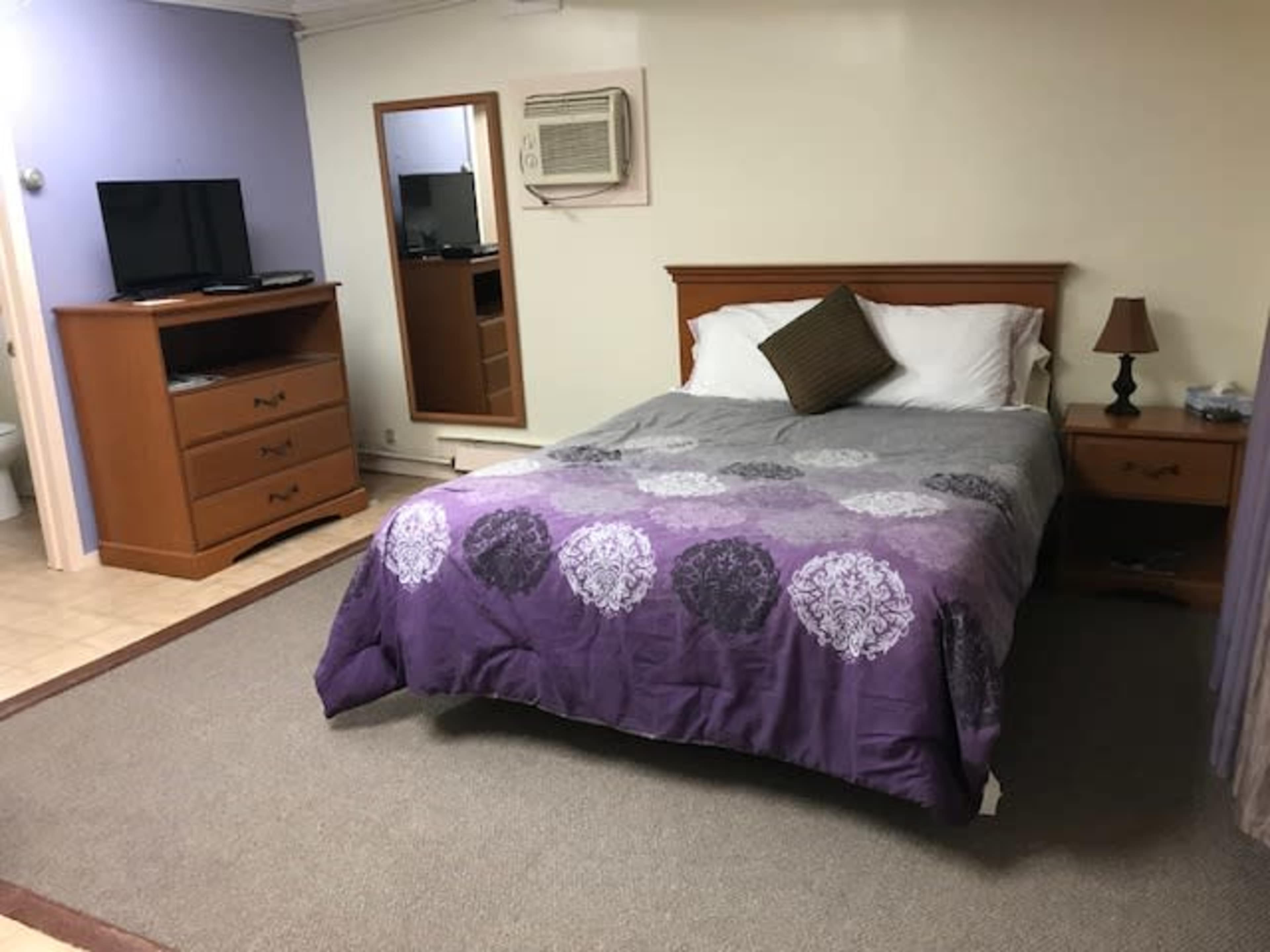 A bedroom features a bed with a purple patterned coverlet, a nightstand with a lamp, a television on a wooden dresser, and a mirror hanging on the wall.