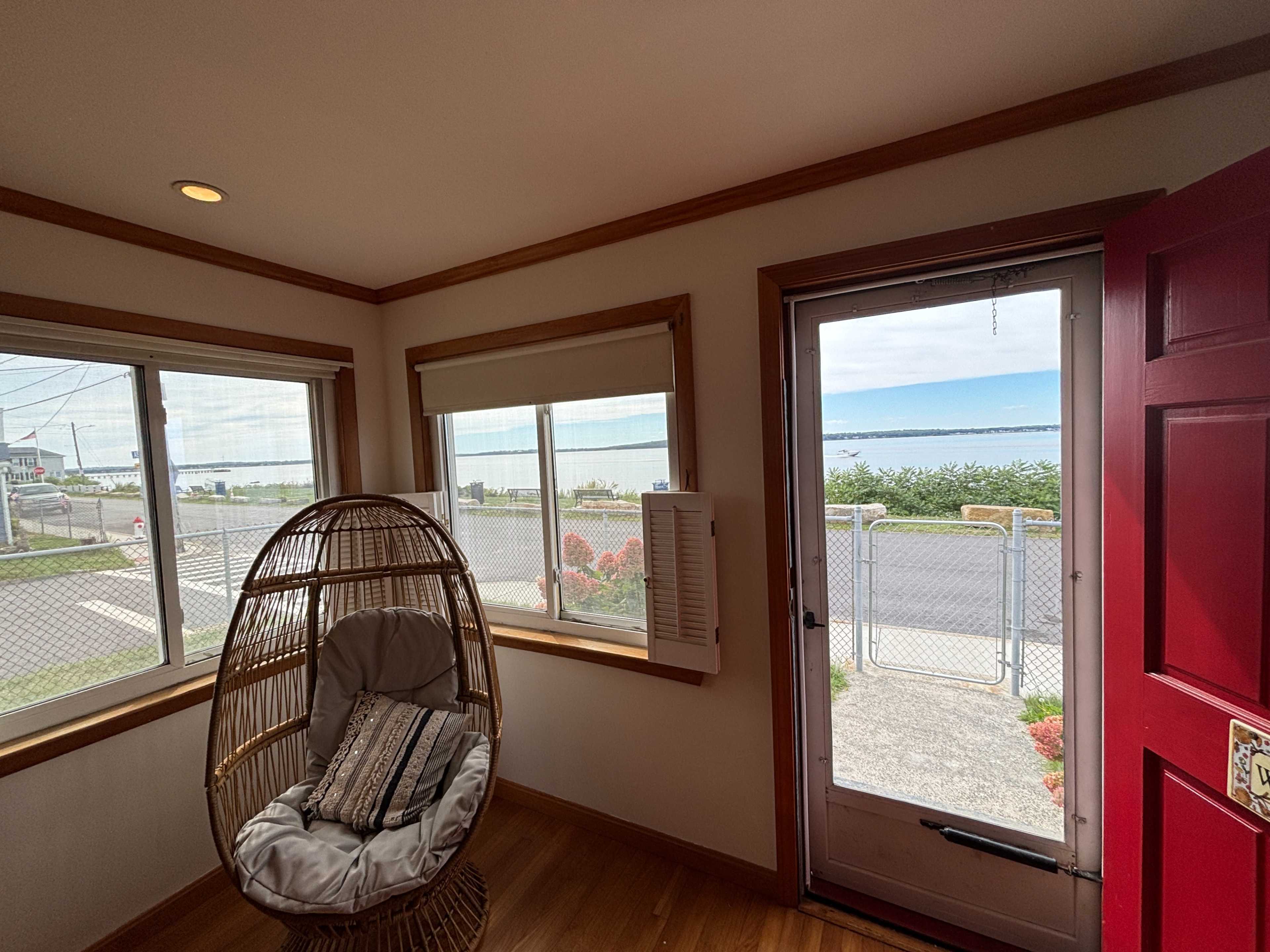 The image shows a bright entryway with a large window facing a body of water, a door with a red frame, and a hanging chair in the corner.
