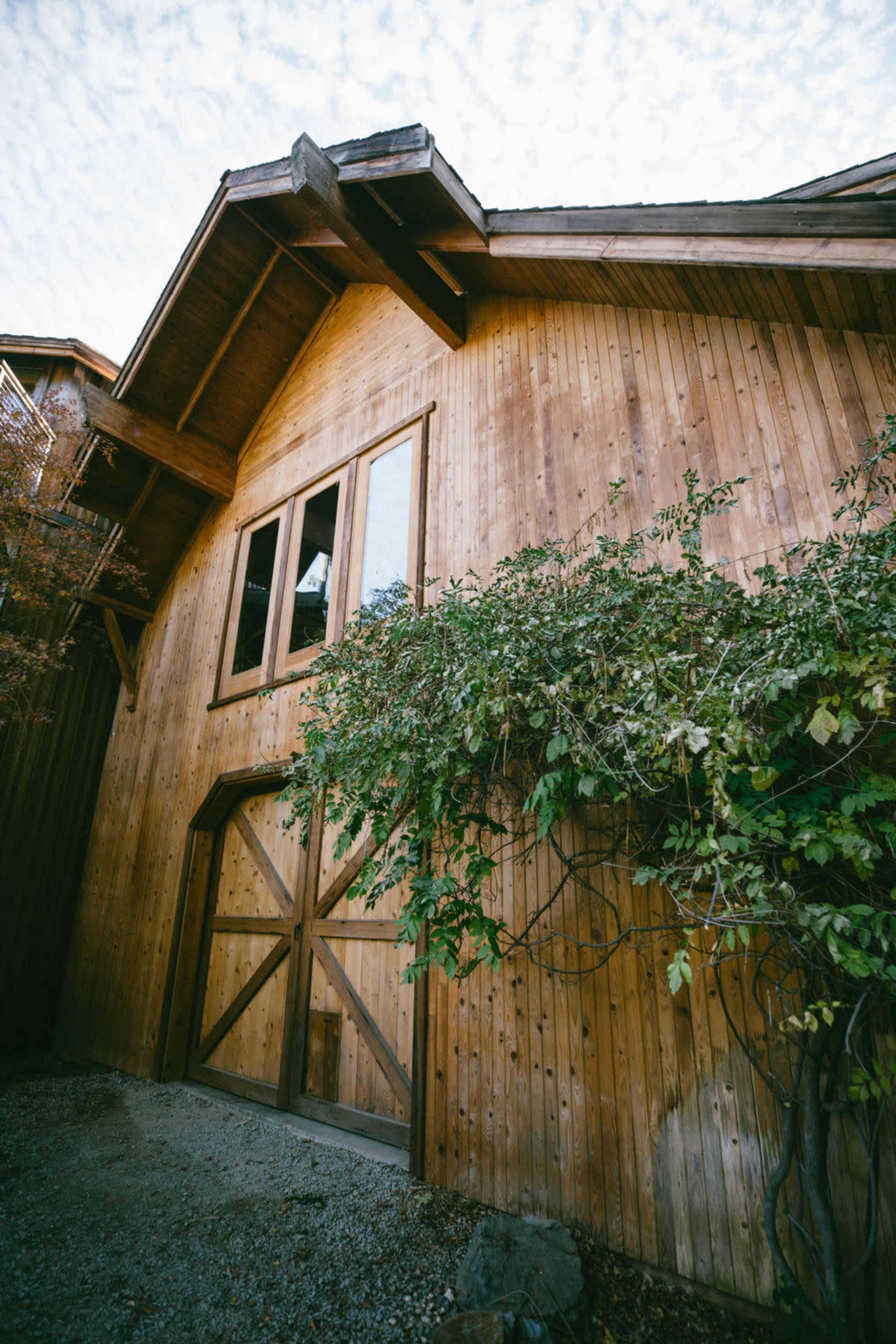 The image shows a wooden barn with a large double door and several windows, surrounded by greenery and gravel.