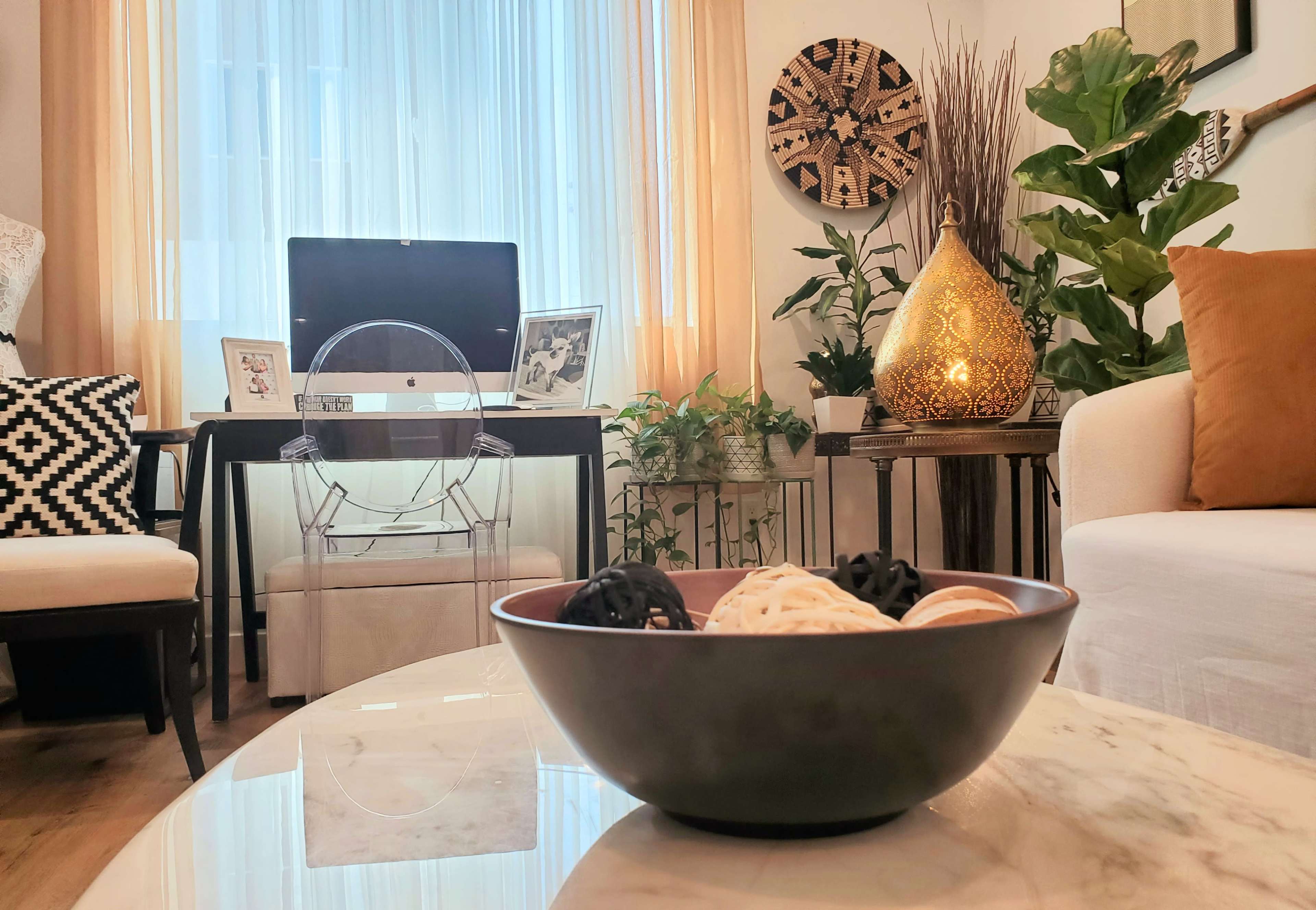 A cozy living room features a decorative bowl of cookies on a marble coffee table, with a computer desk, plants, and an illuminated decorative lamp in the background.