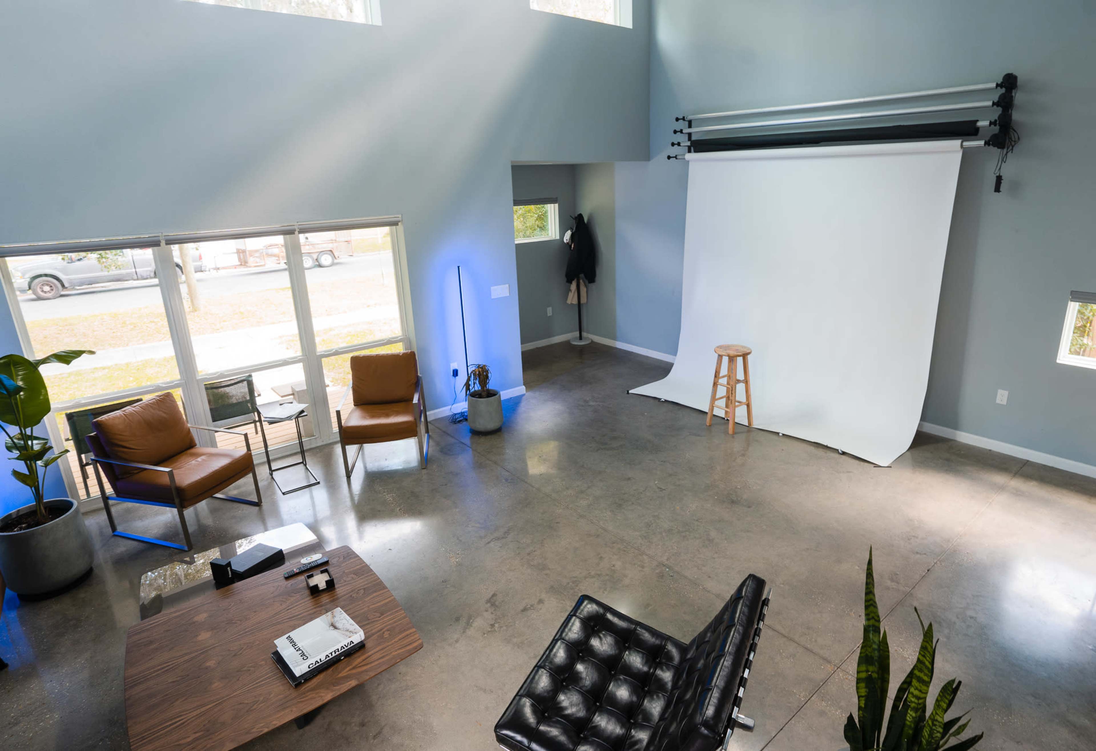 A spacious studio with a white backdrop, a wooden stool, and modern furniture, illuminated by natural light from large windows.