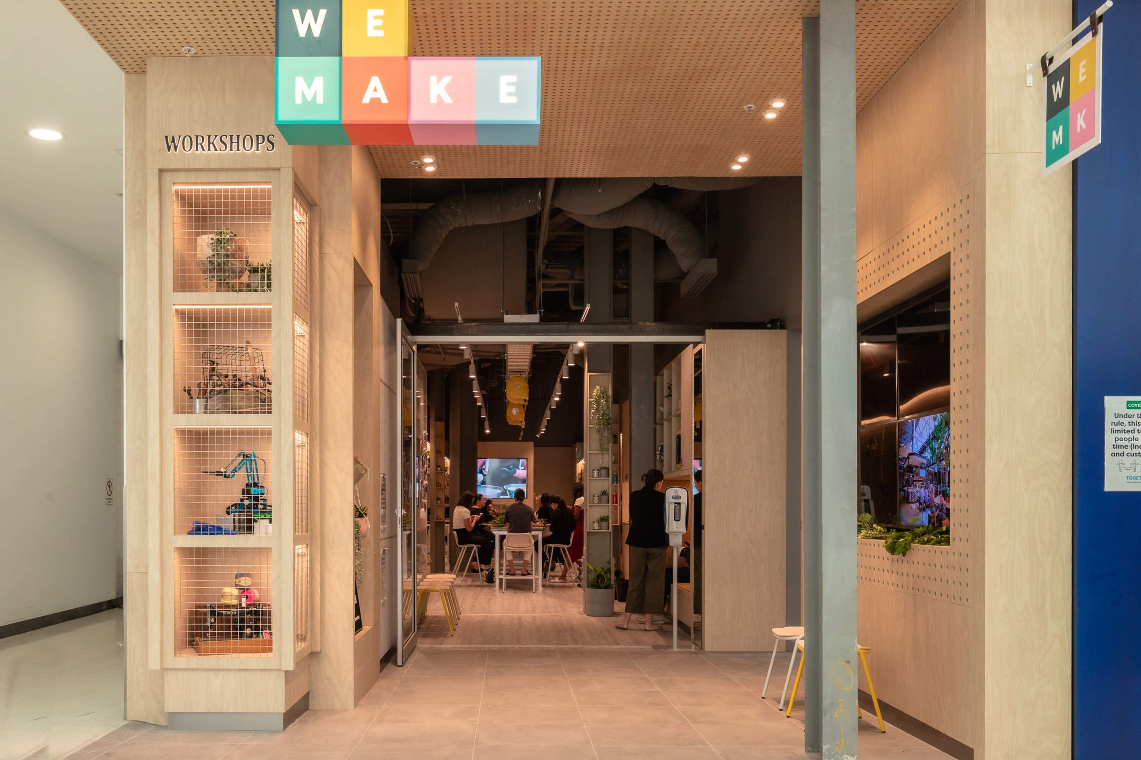 A large, modern entrance to a workshop space labeled "WE MAKE," with shelves displaying various items and a seating area visible in the background.