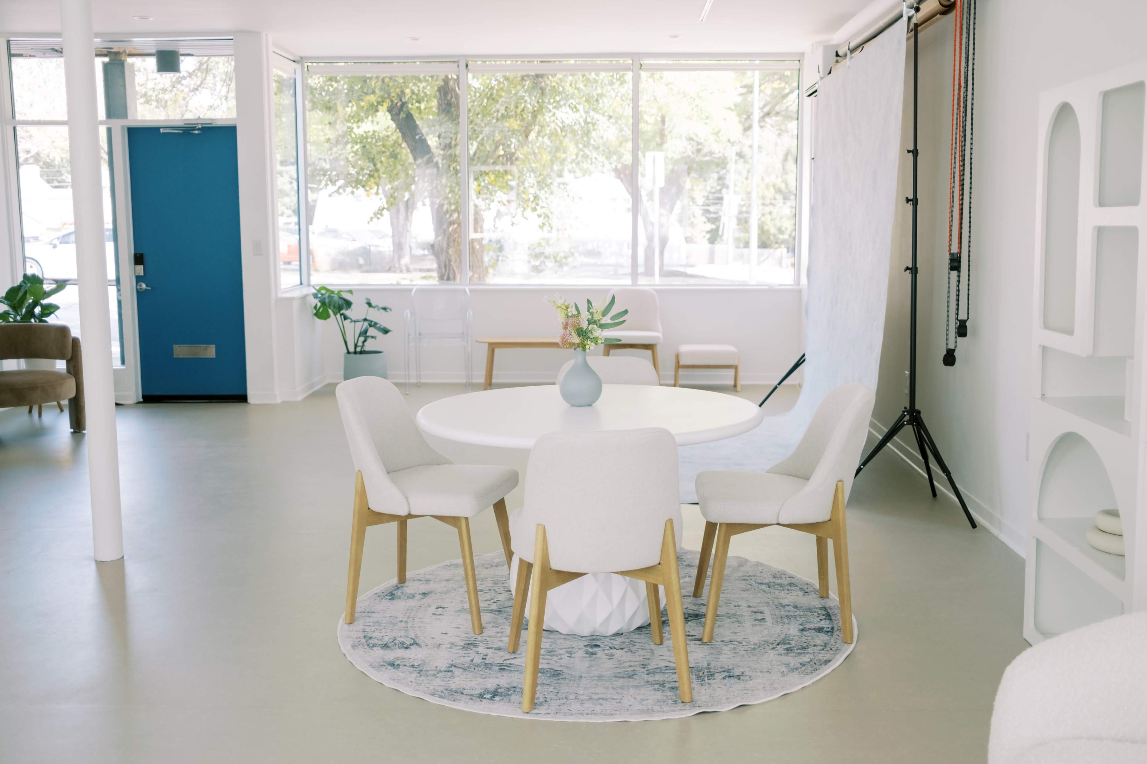 The image shows a light-filled room with a round table surrounded by four upholstered chairs, set against a backdrop of large windows and greenery.