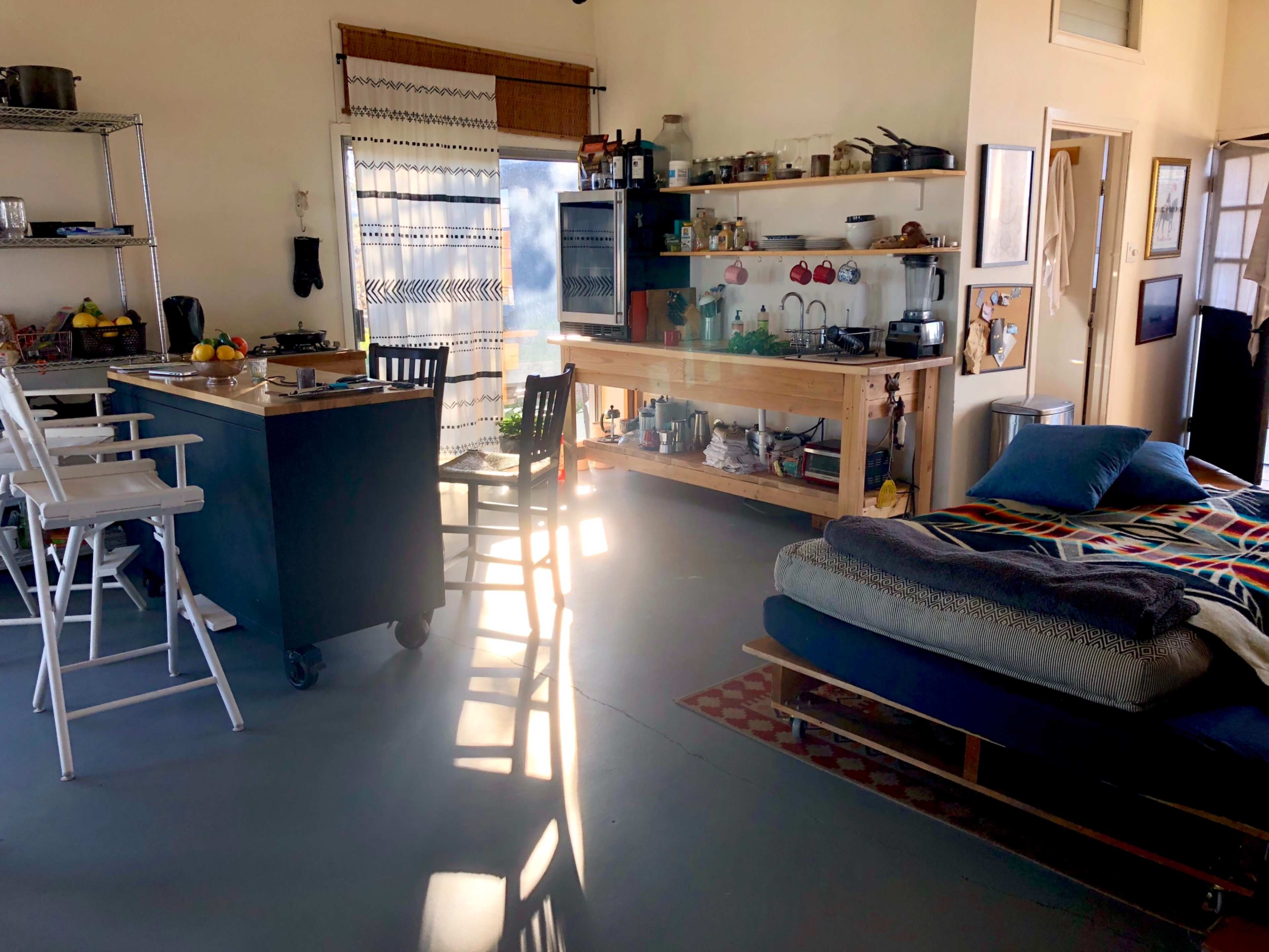 A bright and spacious open living area with a kitchen island, a dining table with high chairs, and a cozy seating area featuring a couch and a patterned throw.