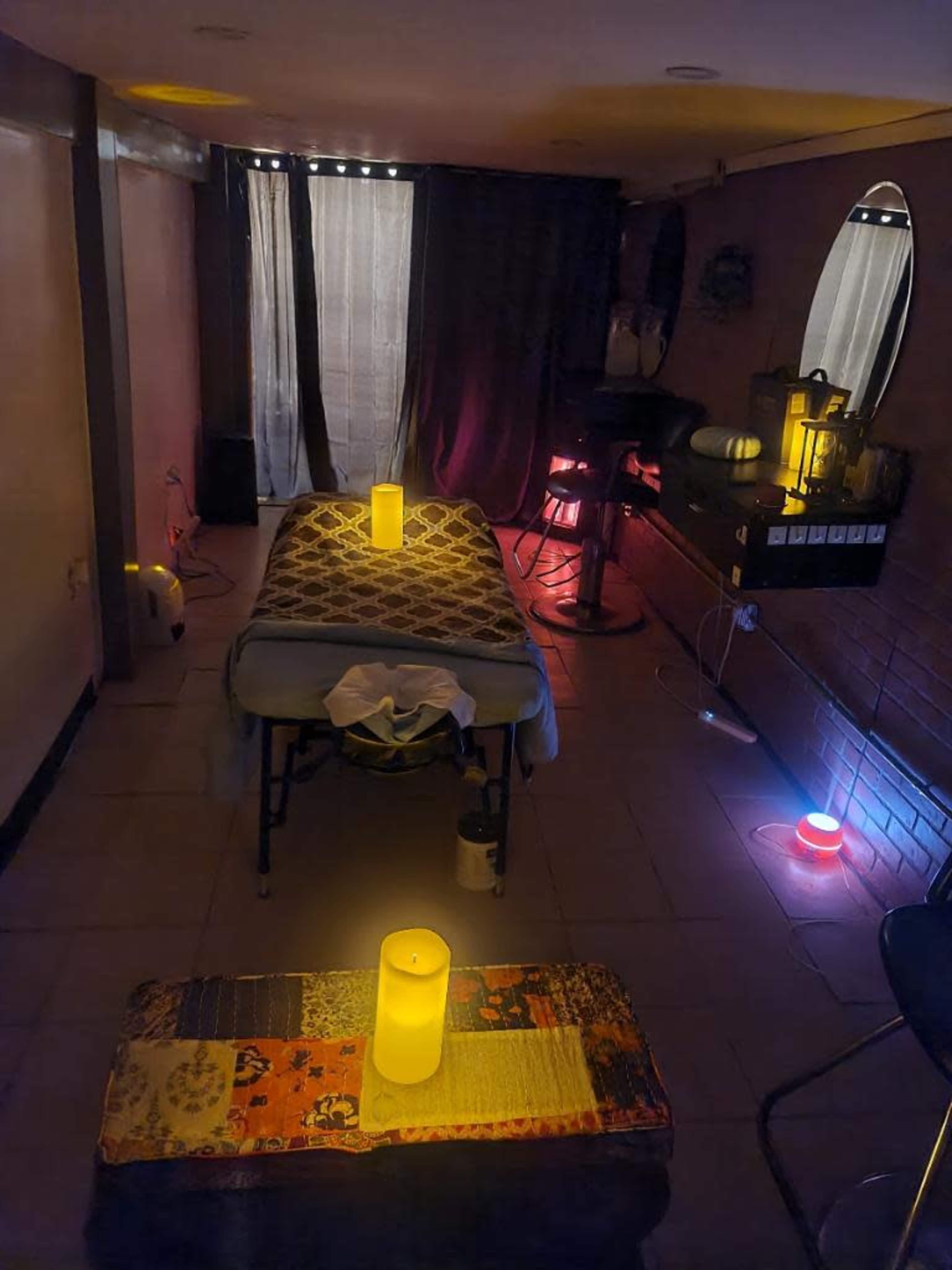 A dimly lit room features a massage table in the center, surrounded by candles and colored lights against the walls.