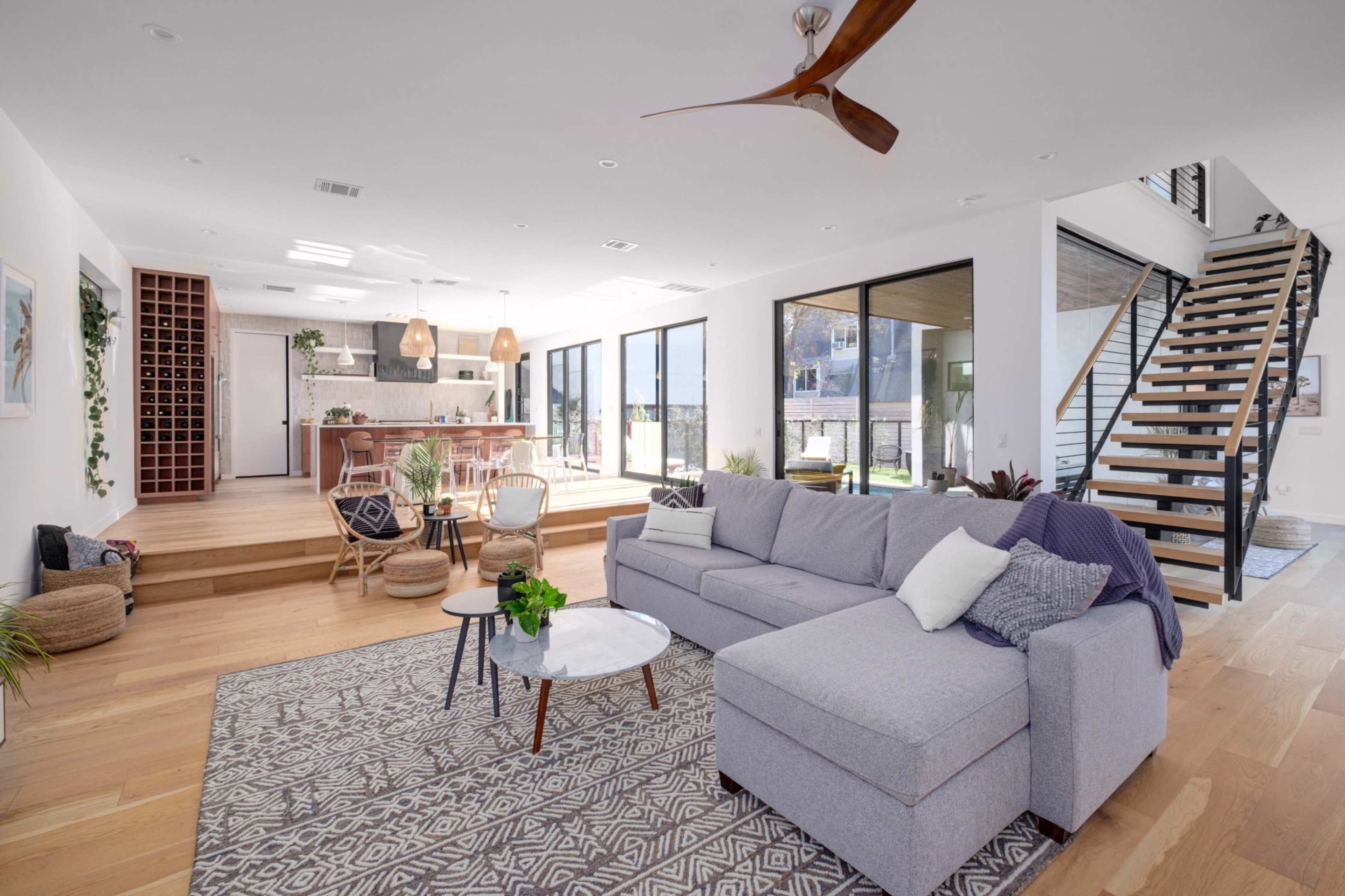 A modern living room with a gray sectional sofa, a round white coffee table, and large windows that open to an outdoor area, complemented by wooden flooring and a staircase in the background.