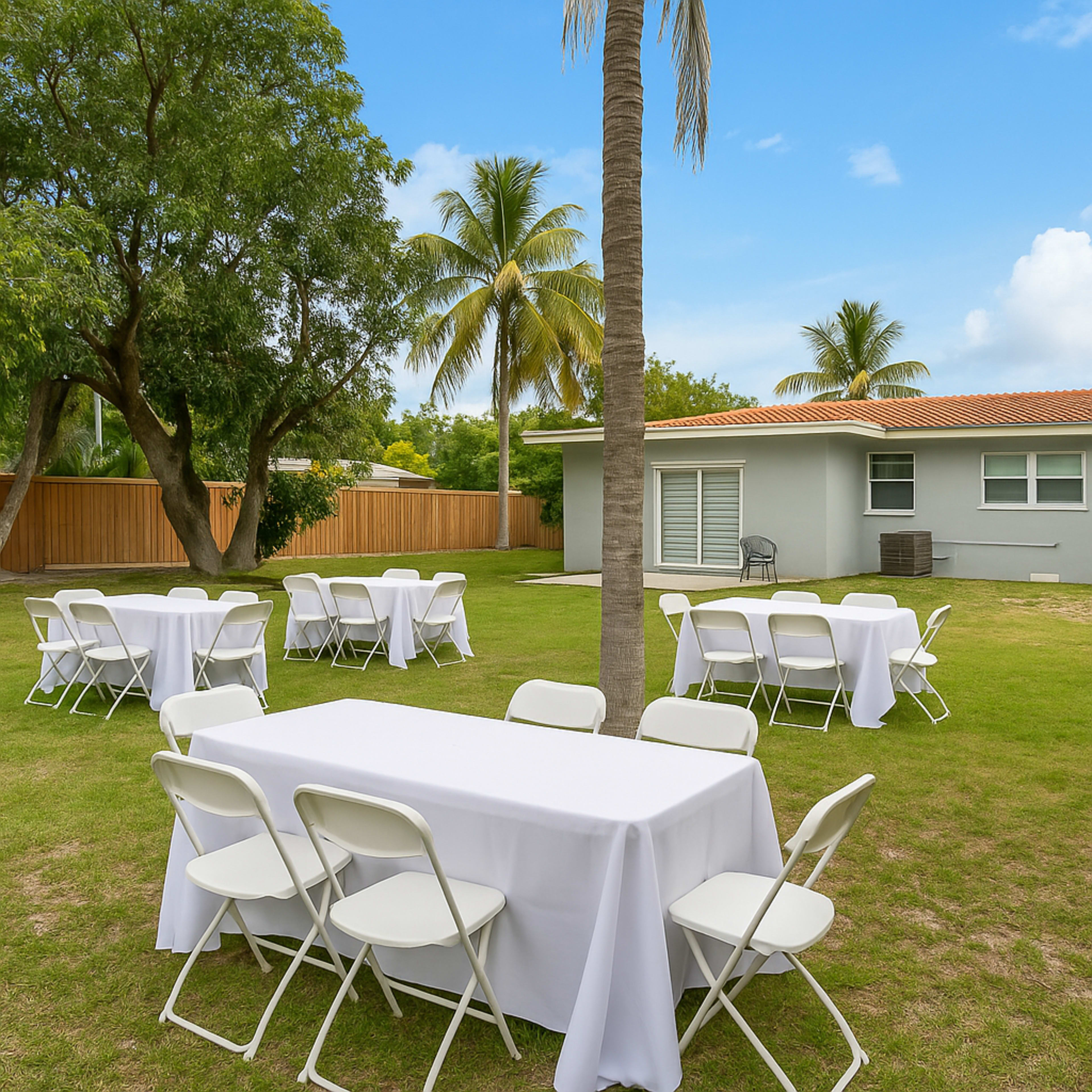 The image shows an outdoor setting with several tables covered in white cloth and surrounded by folding chairs, set on a grassy area with palm trees and a gray house in the background.