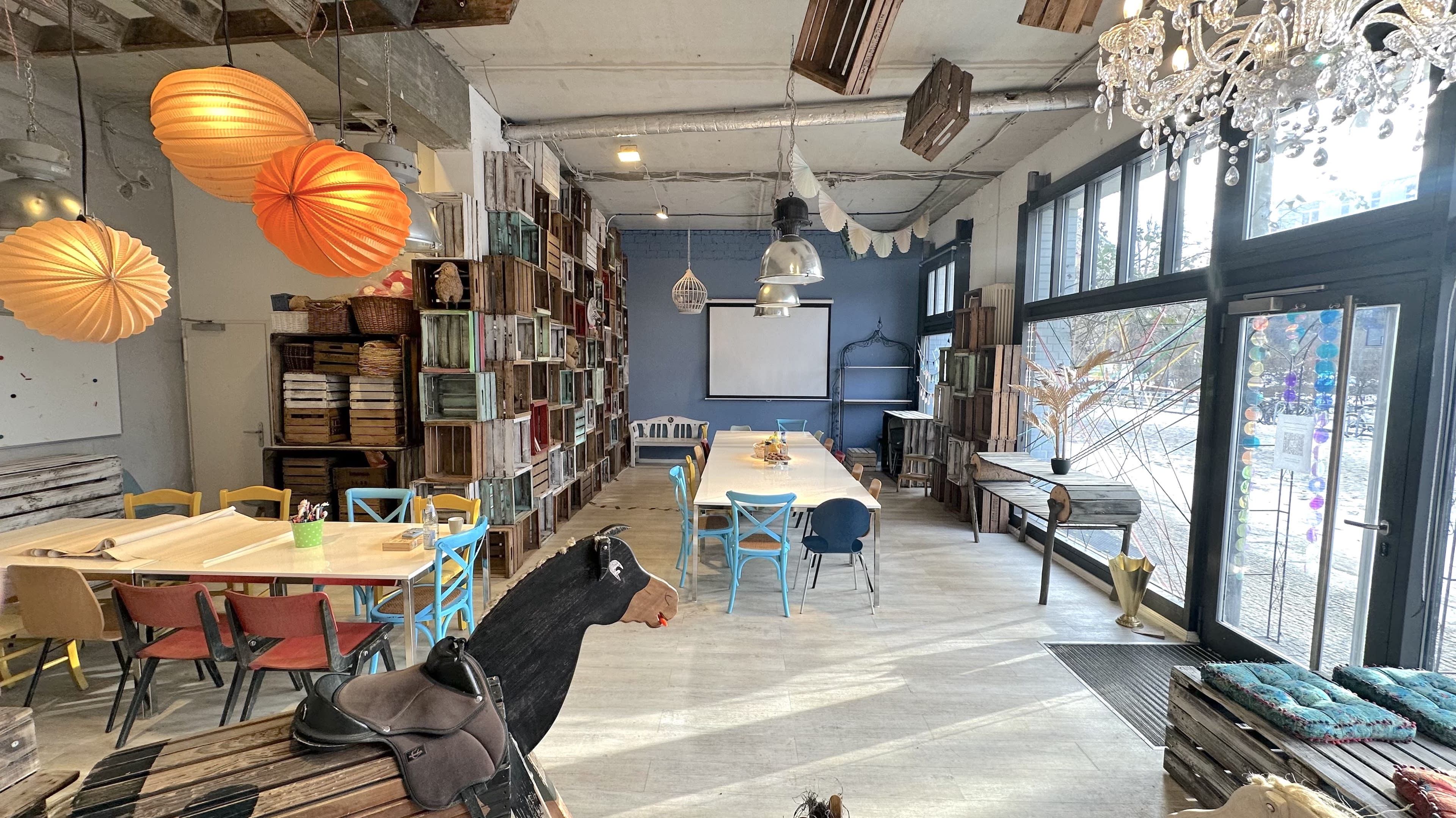 The image shows a brightly lit creative workspace featuring a long table surrounded by colorful chairs, with shelves filled with various objects and decorative elements, including hanging light fixtures.