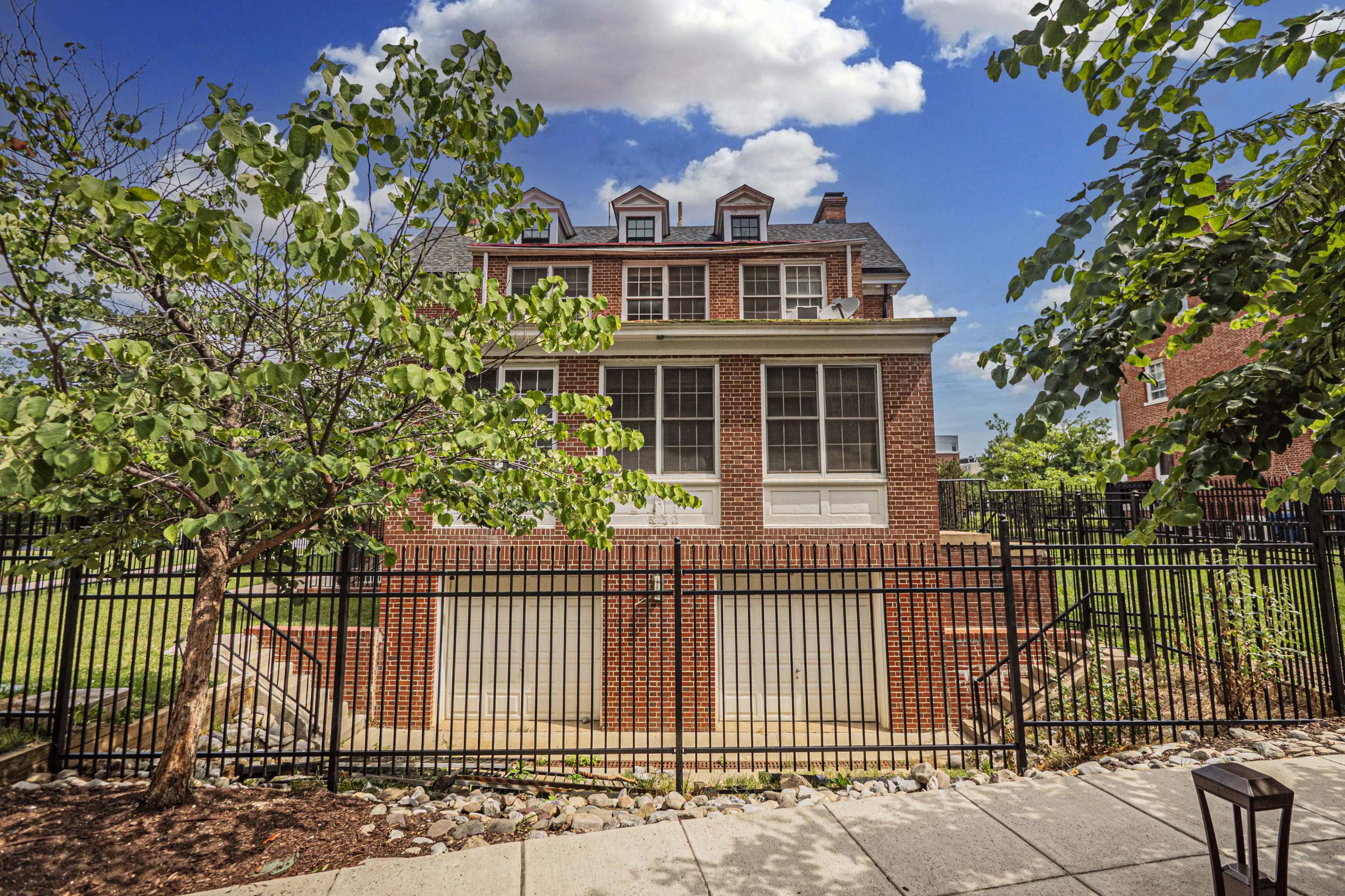 The image shows a brick house with multiple gabled windows, situated behind a black metal fence, surrounded by greenery under a partly cloudy sky.