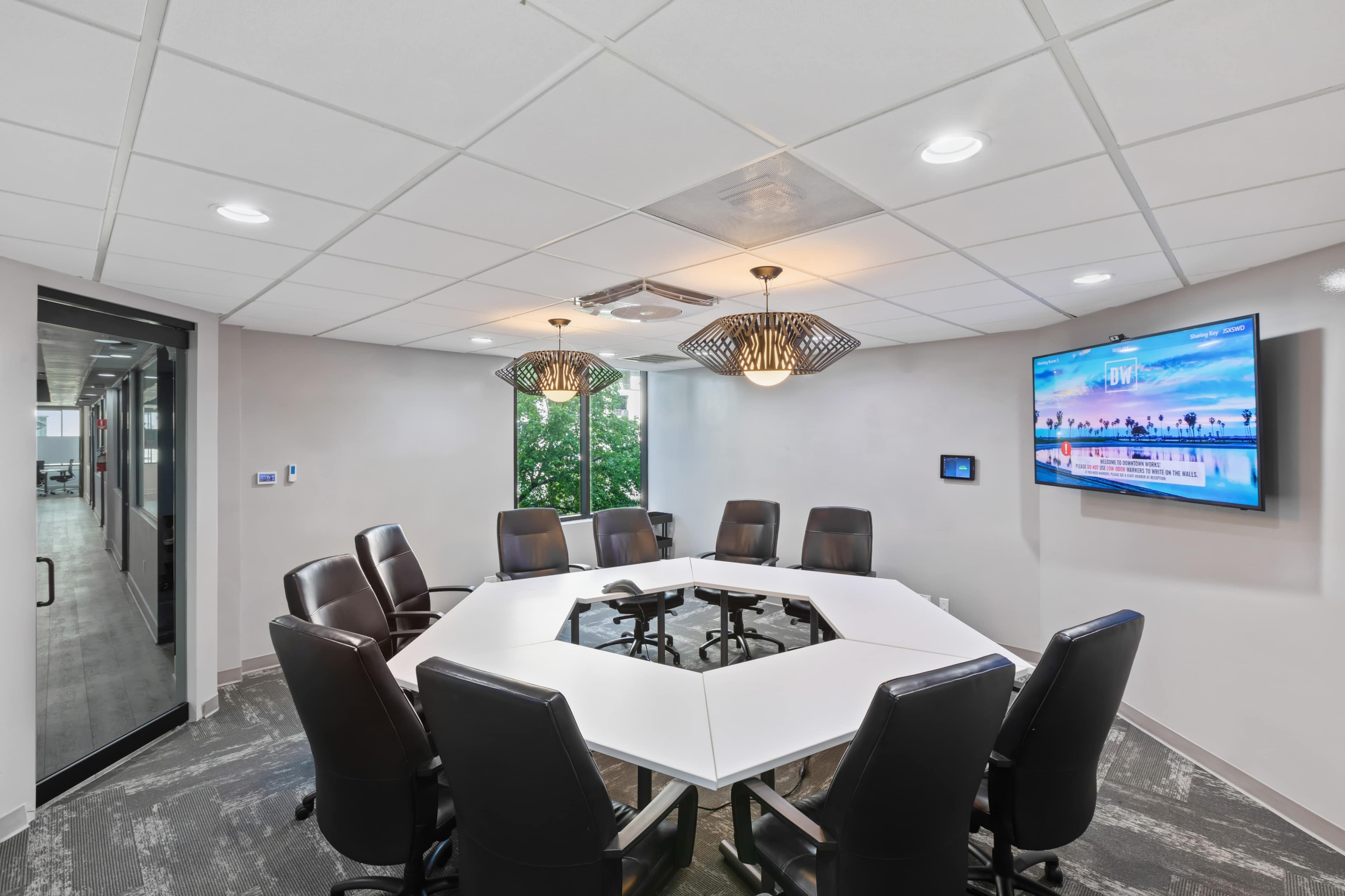 A modern conference room with a large octagonal table surrounded by black leather chairs and a wall-mounted television displaying a scenic image.