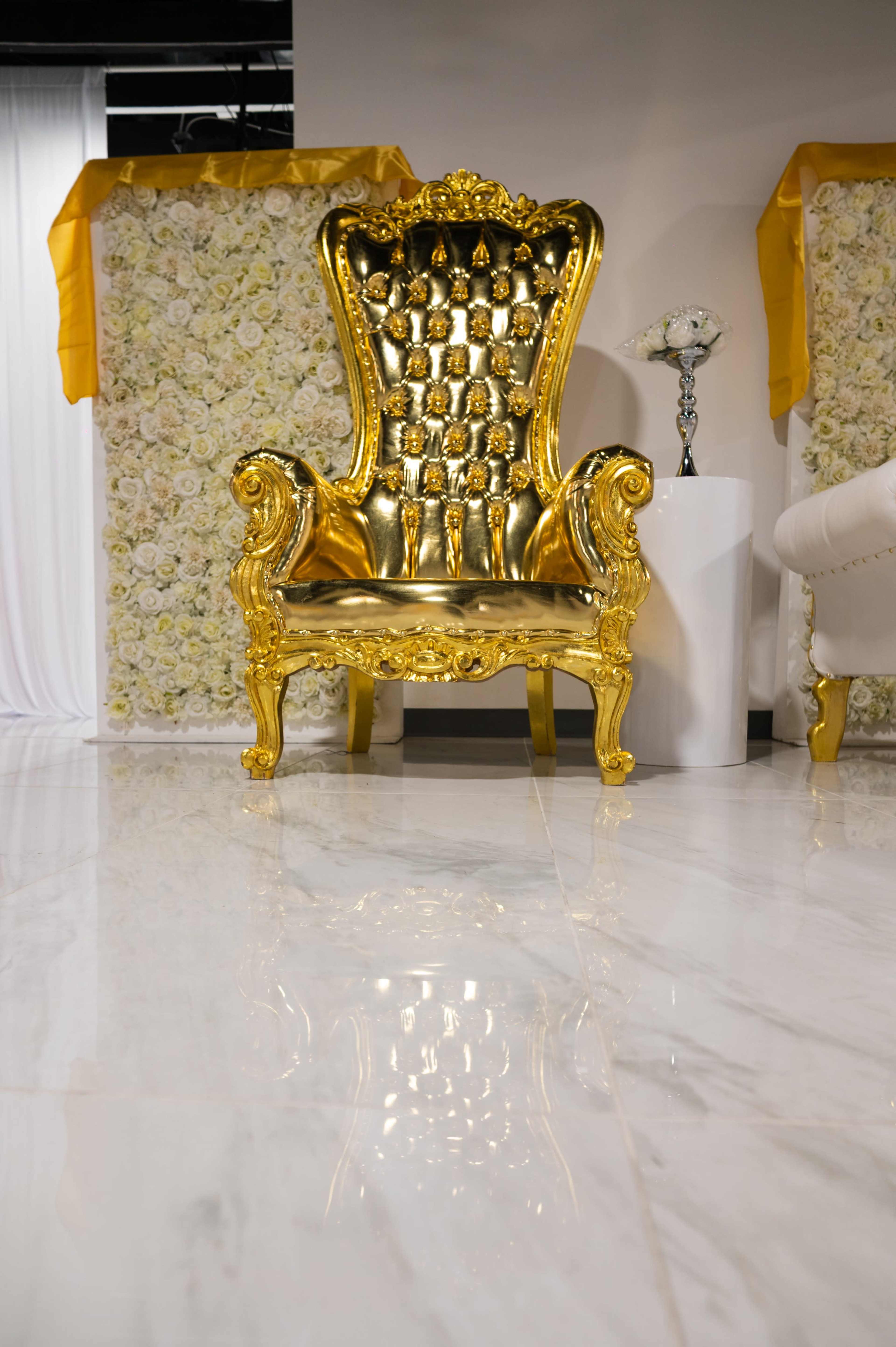 The image shows a luxurious gold throne chair in an elegantly decorated event space with floral backdrops and polished marble flooring.