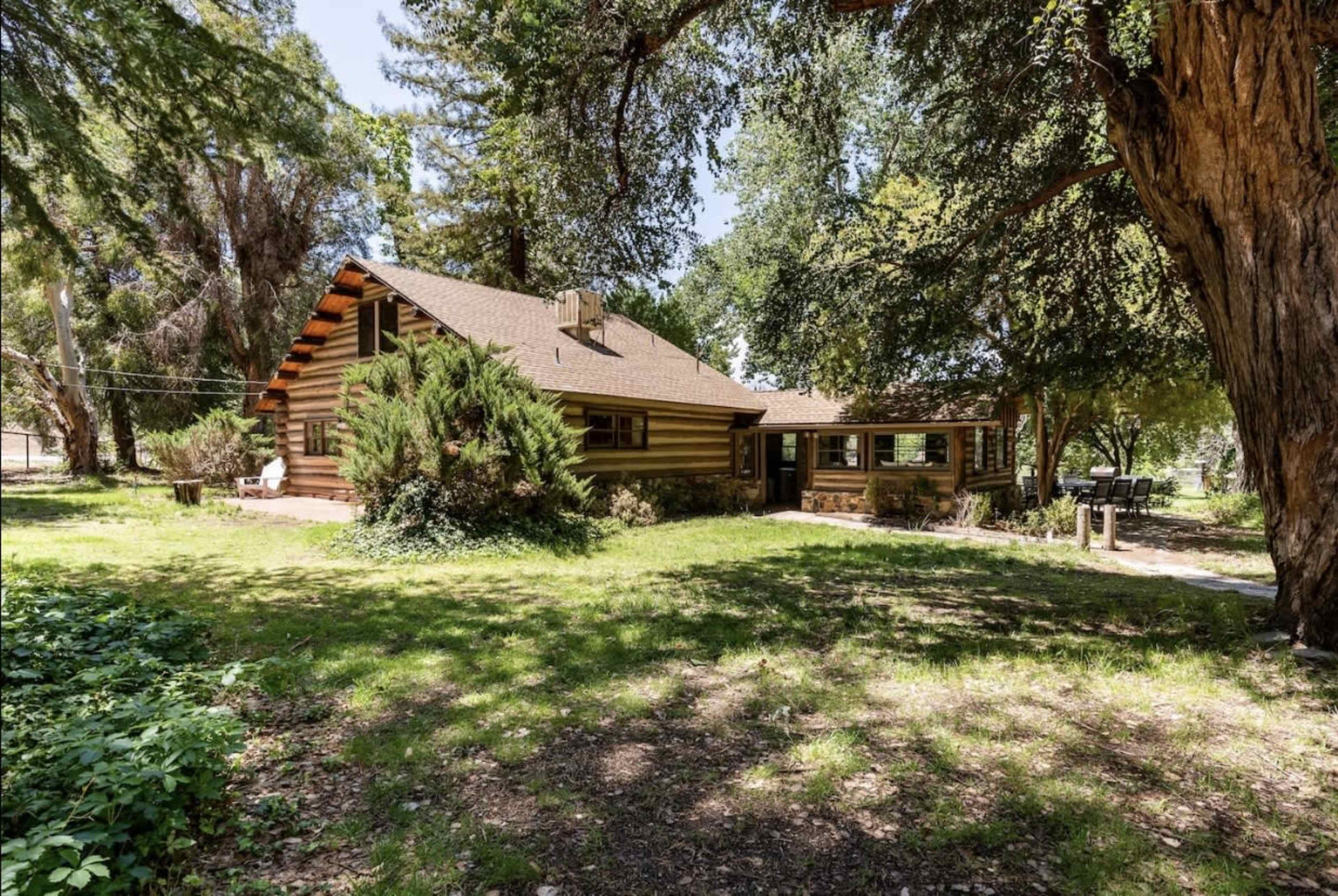 A rustic log cabin with a sloped roof sits among green trees and grassy land.