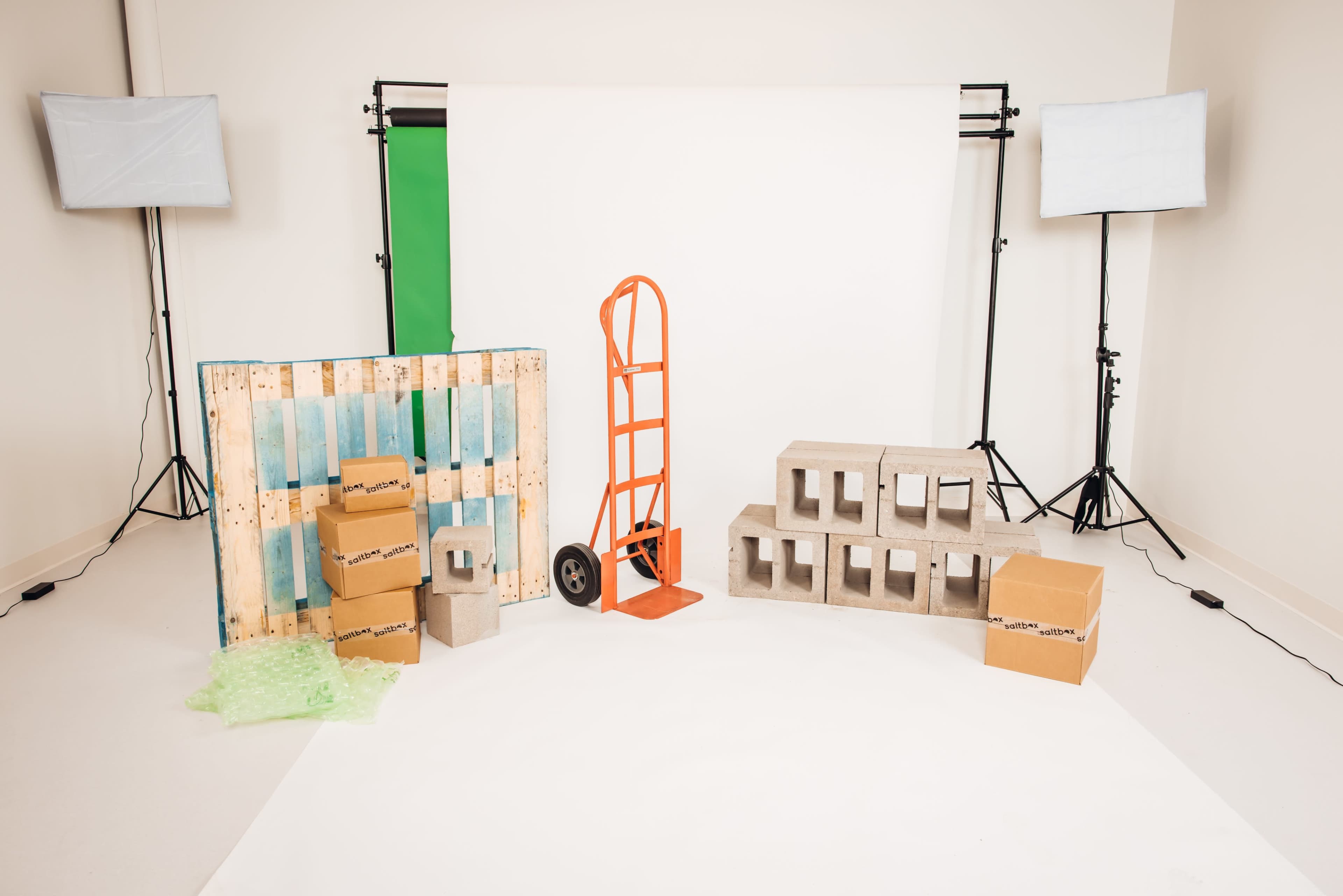 The scene features a studio setup with a green backdrop, several boxes, a hand truck, and concrete blocks arranged on a white floor, illuminated by two soft box lights.
