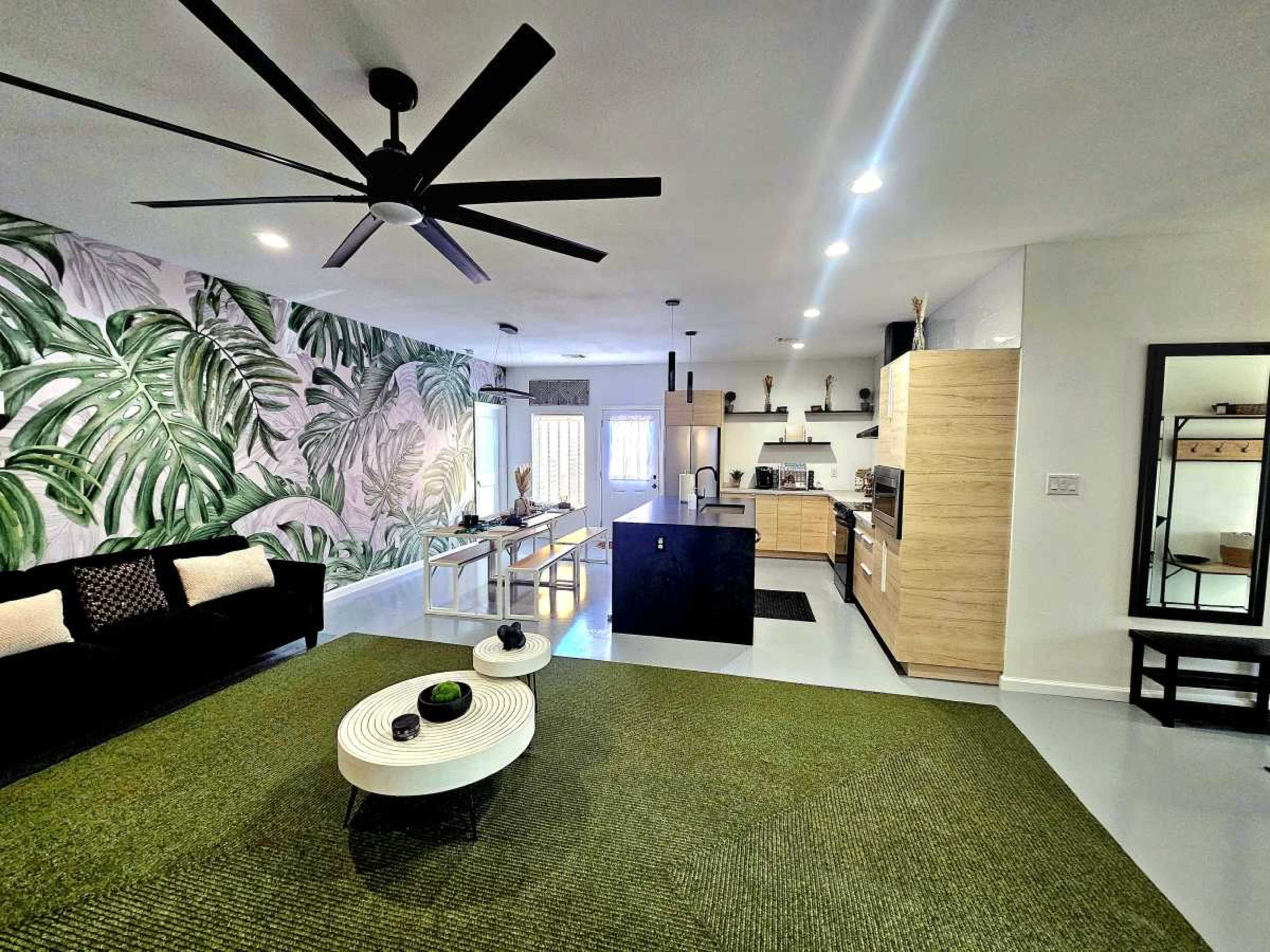 The image shows a modern living space with a large area rug, a black sofa, a dining table, a kitchen, and a tropical-themed accent wall.