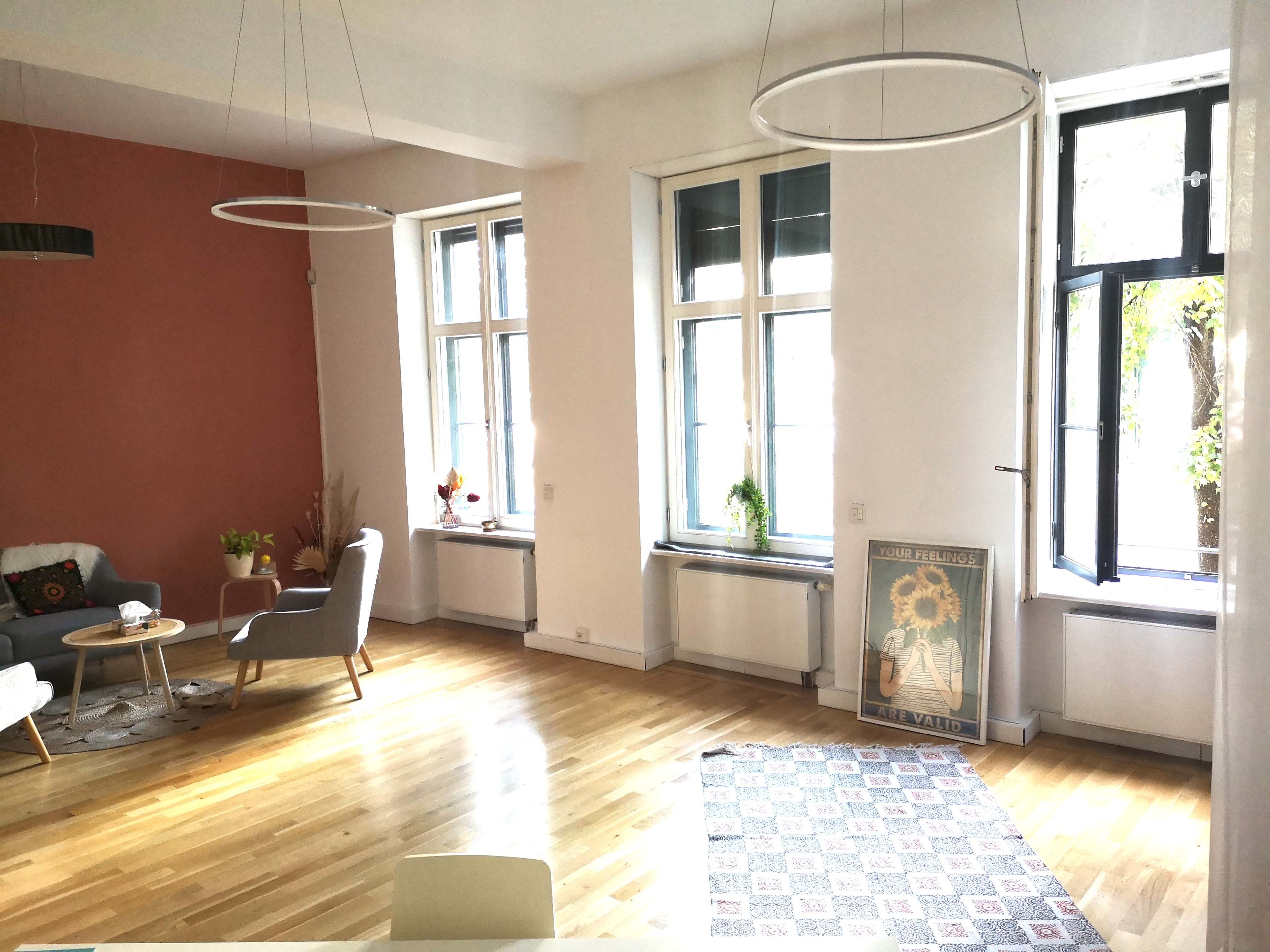 The image shows a modern apartment living area featuring a light wood floor, large windows with open shutters, and minimalistic furniture including a gray sofa and a small round table.