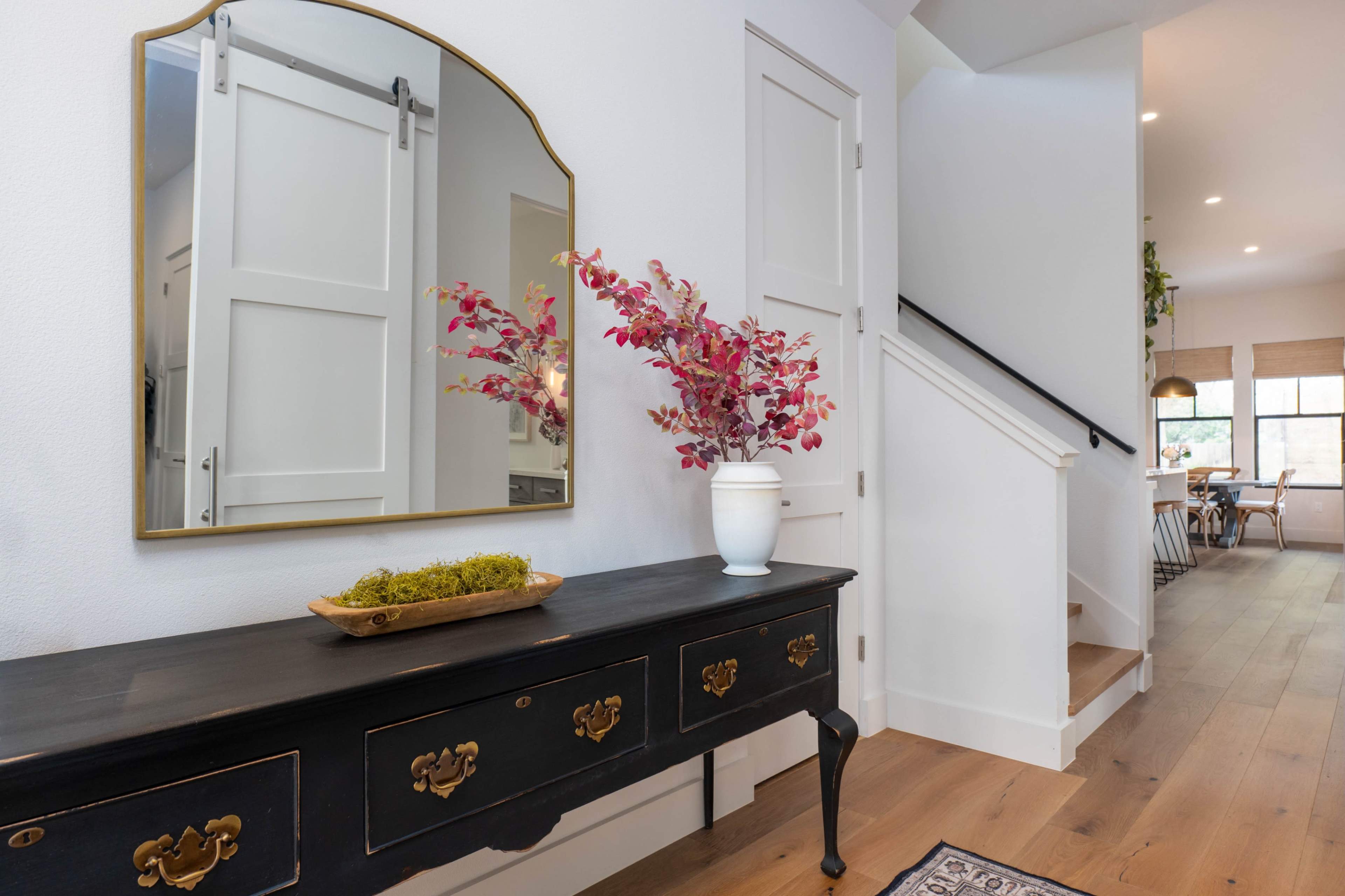 A hallway features a black console table with a decorative vase of flowers, a large mirror, and stairs leading to the upper level.