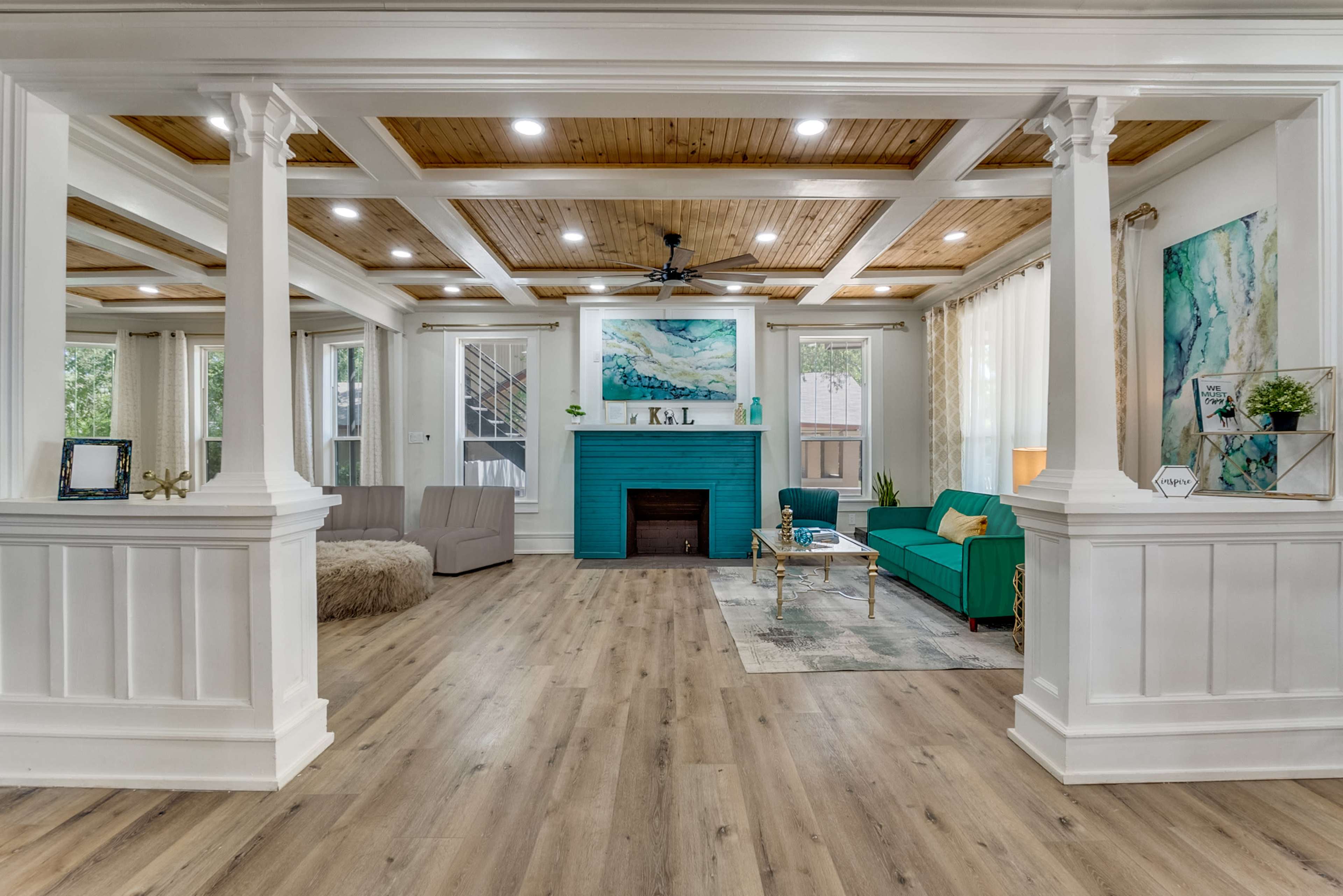 The image shows a spacious living room with wooden beams on the ceiling, white pillars, and teal furniture, featuring a fireplace and large windows.