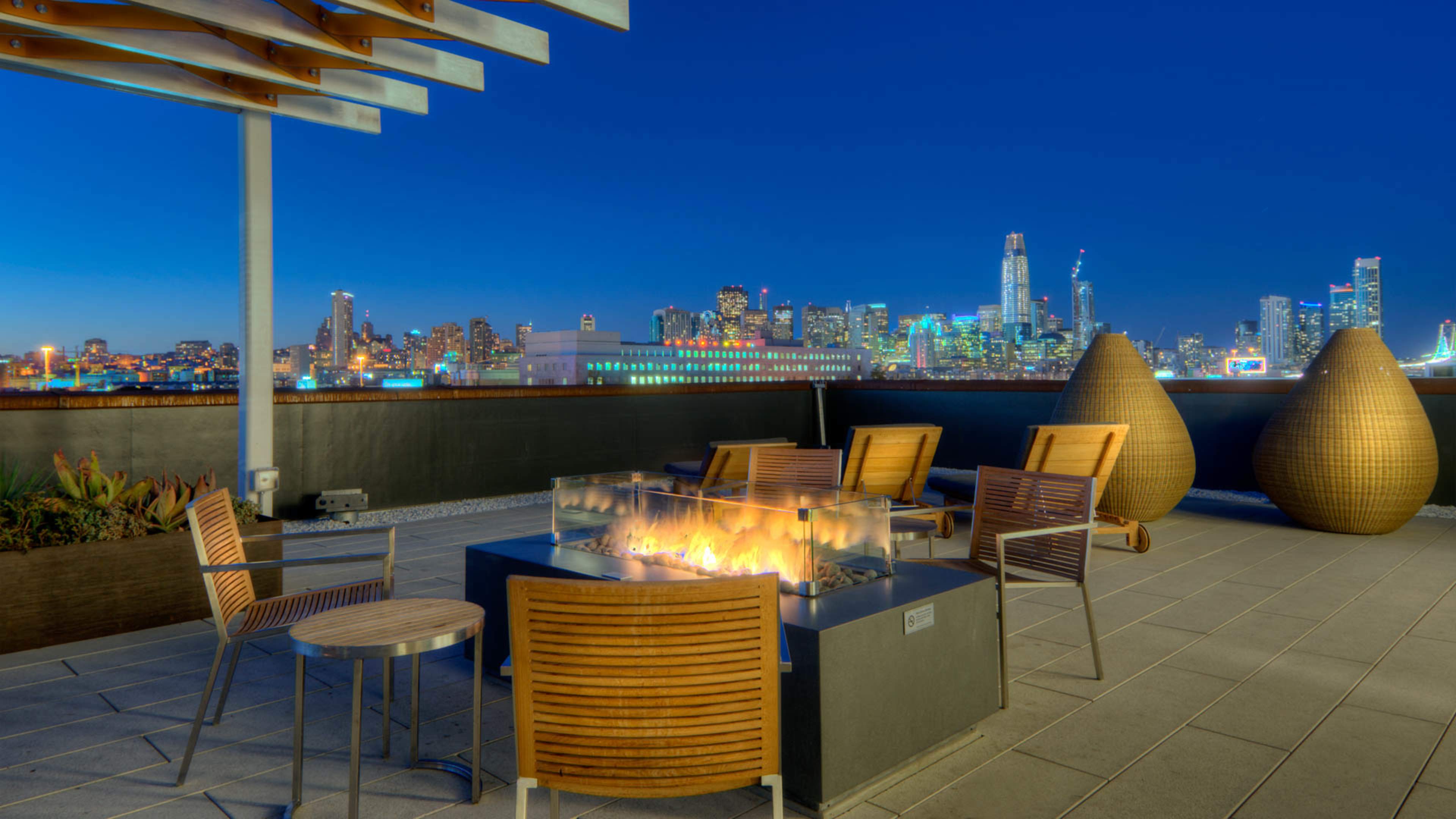 A rooftop terrace features a fire pit surrounded by wooden chairs, with a city skyline illuminated by night in the background.