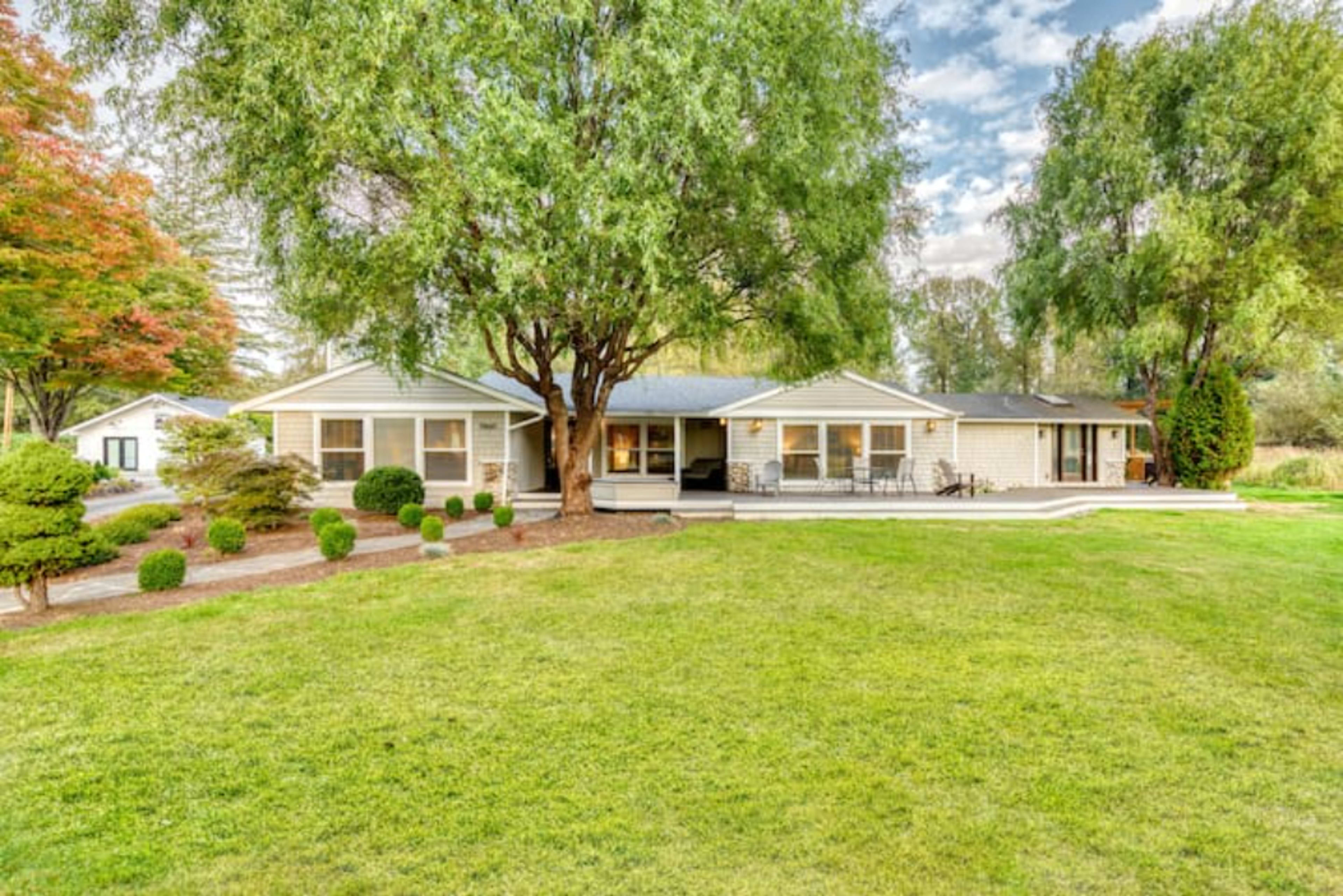A single-story house with a large lawn, surrounded by trees and shrubs, features a patio area in the backyard.