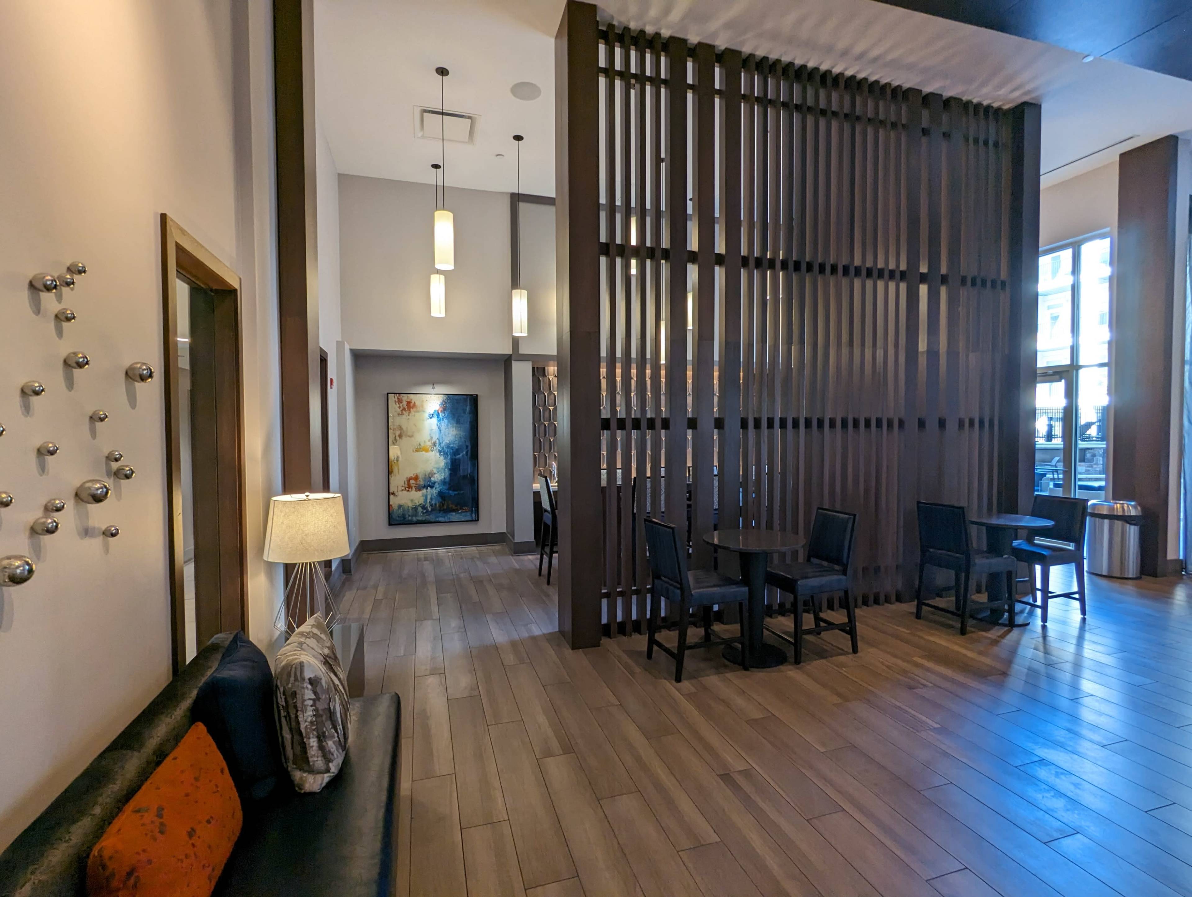 A spacious lobby area with wooden floors, a modern art piece on the wall, and a seating arrangement consisting of black chairs and a small table near a lamp.