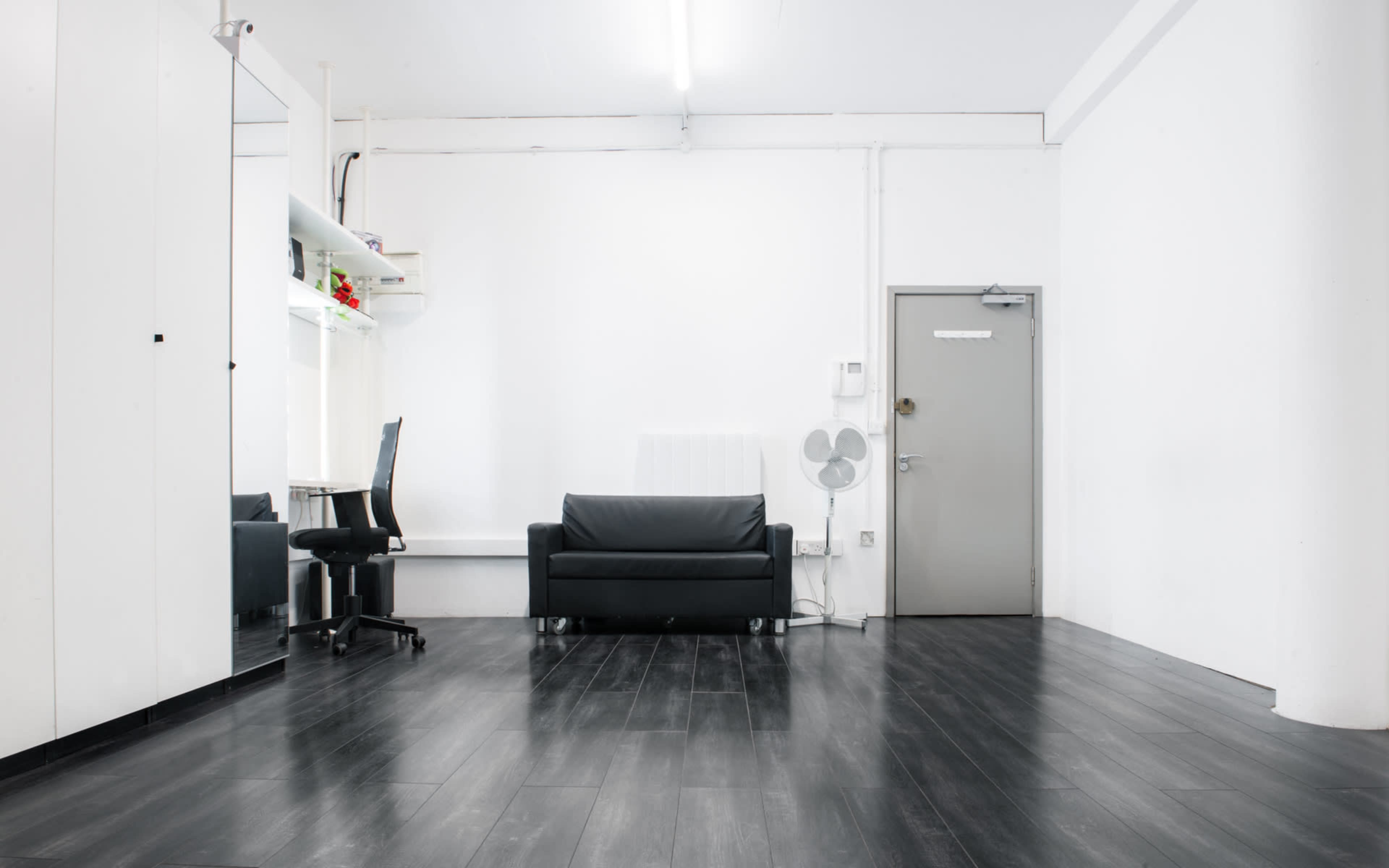 A minimalistic interior featuring a black sofa, a black office chair, and a door, all set against white walls and a dark wooden floor.