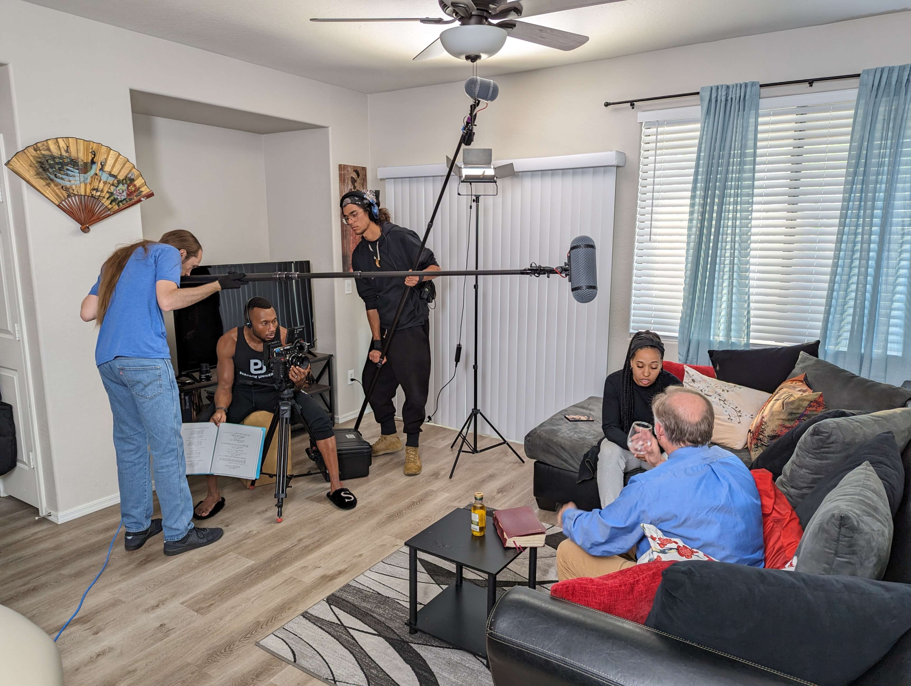 A film crew is recording a scene in a living room, featuring two actors seated on a couch while a sound technician holds a microphone overhead and a camera operator adjusts equipment.
