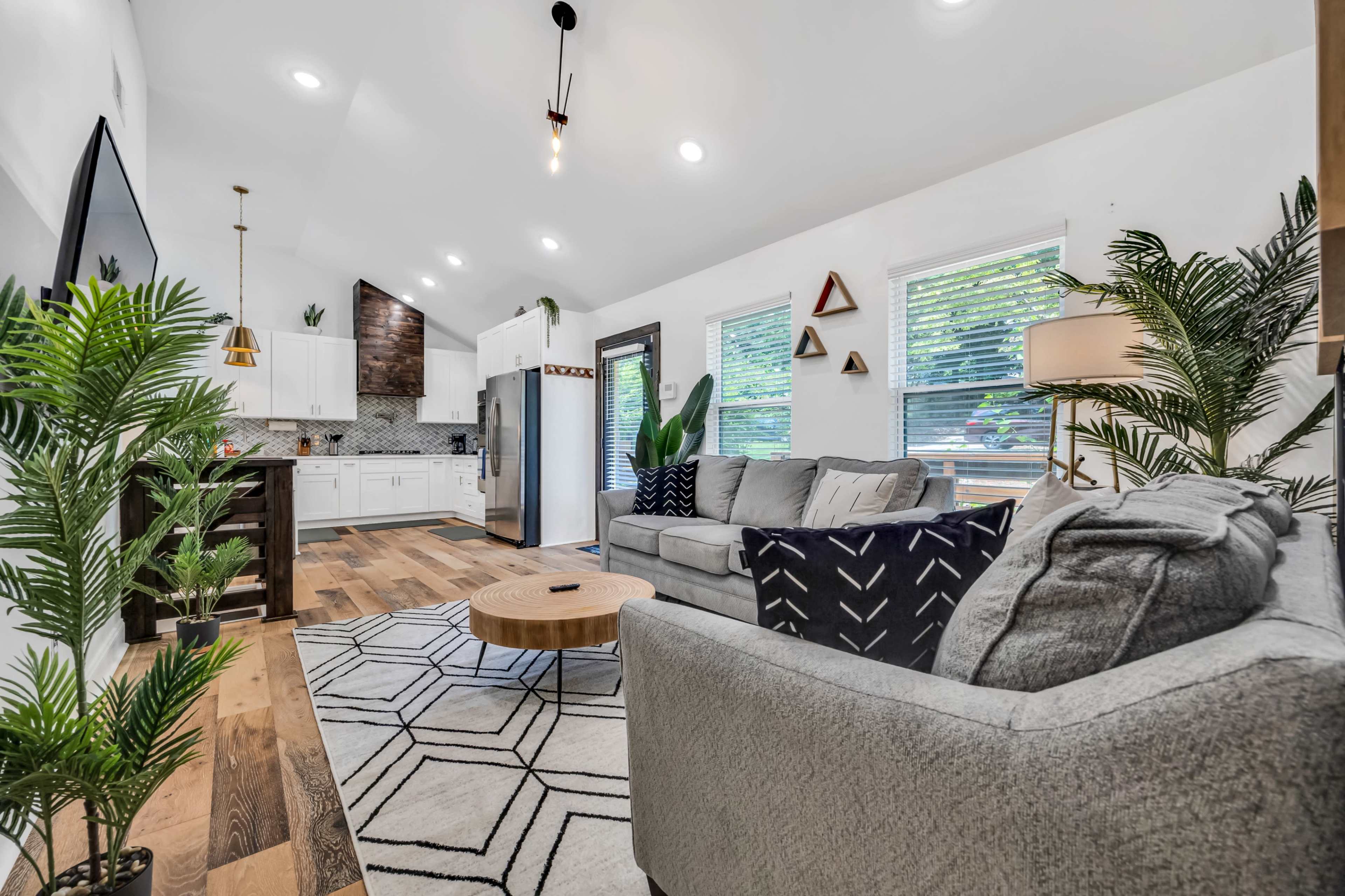 A living room features a gray sectional sofa, a round coffee table, and a kitchen area with white cabinets and a hexagonal backsplash.