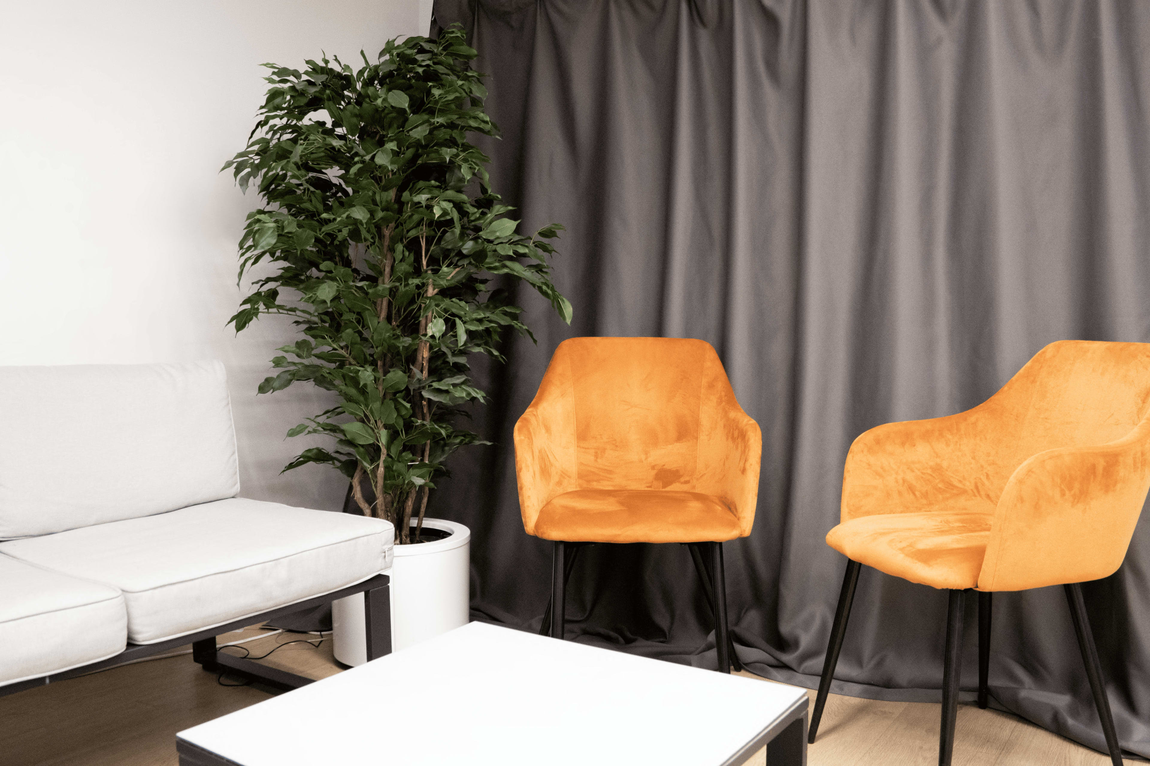 The image shows a modern seating area featuring a light gray sofa, two orange chairs, a potted plant, and a white coffee table in front of a dark gray curtain.