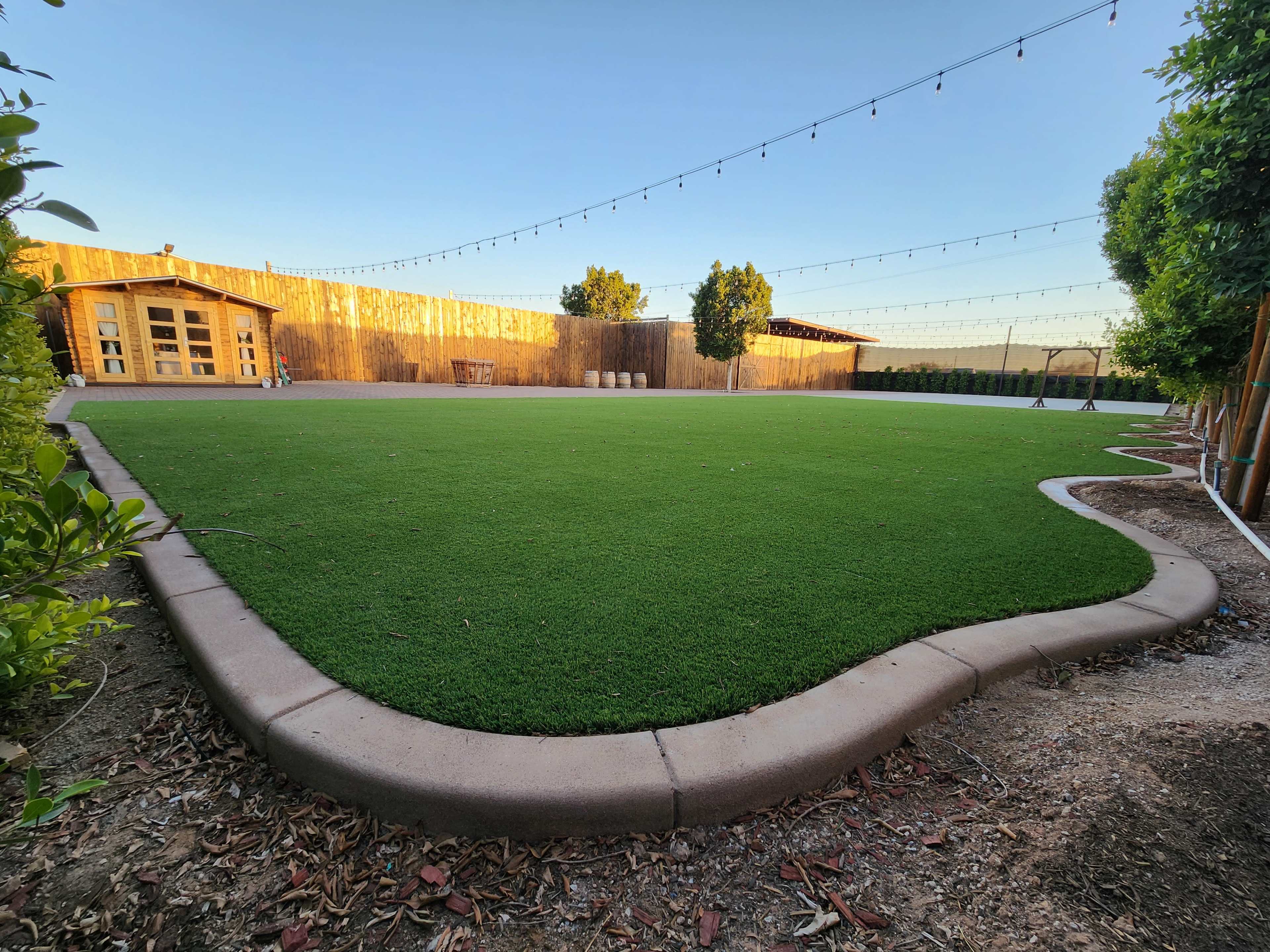 A spacious green lawn surrounded by wooden fences and string lights, with two structures in the background.