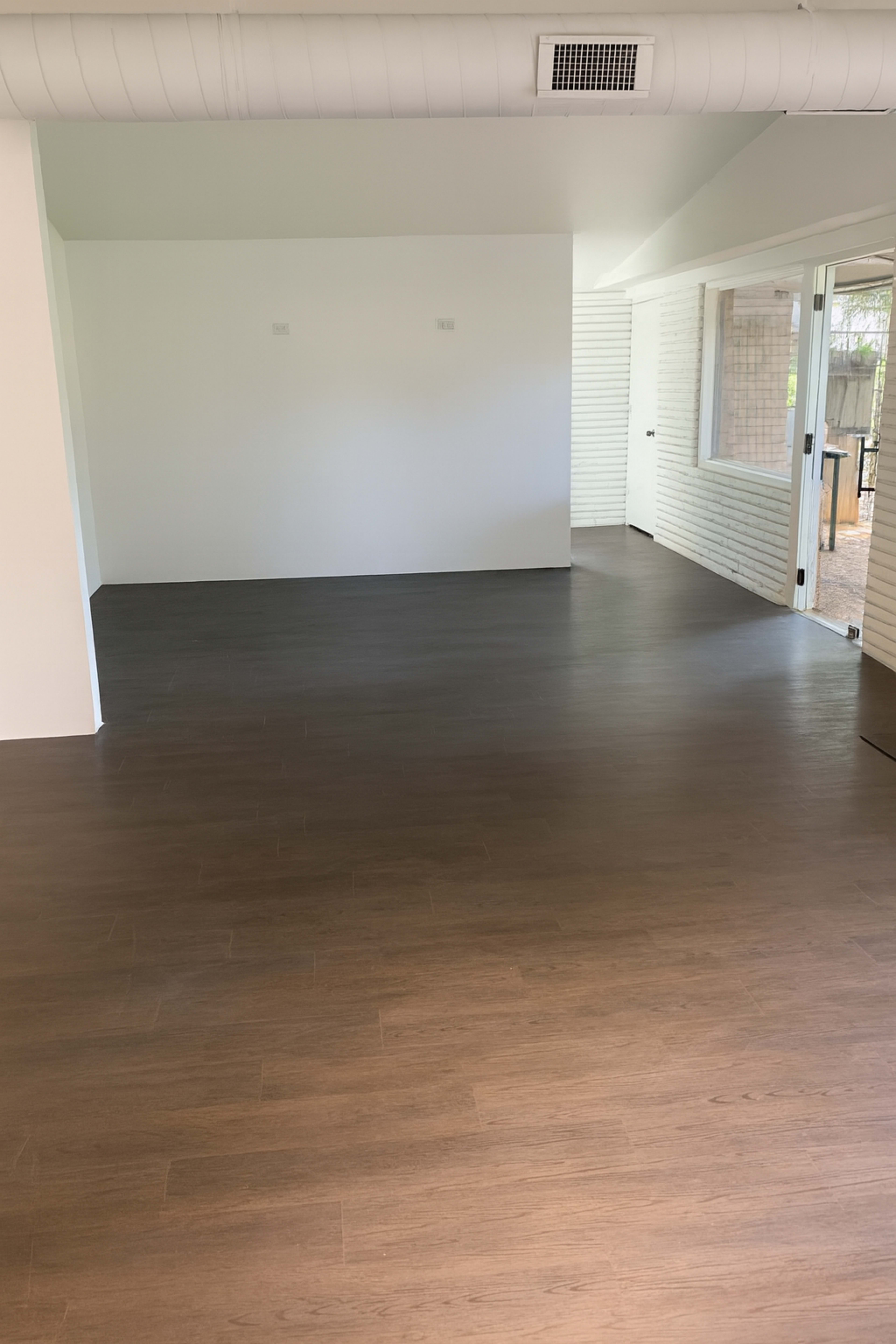 The image shows a spacious room with brown wooden flooring and white walls, featuring large windows that provide natural light.