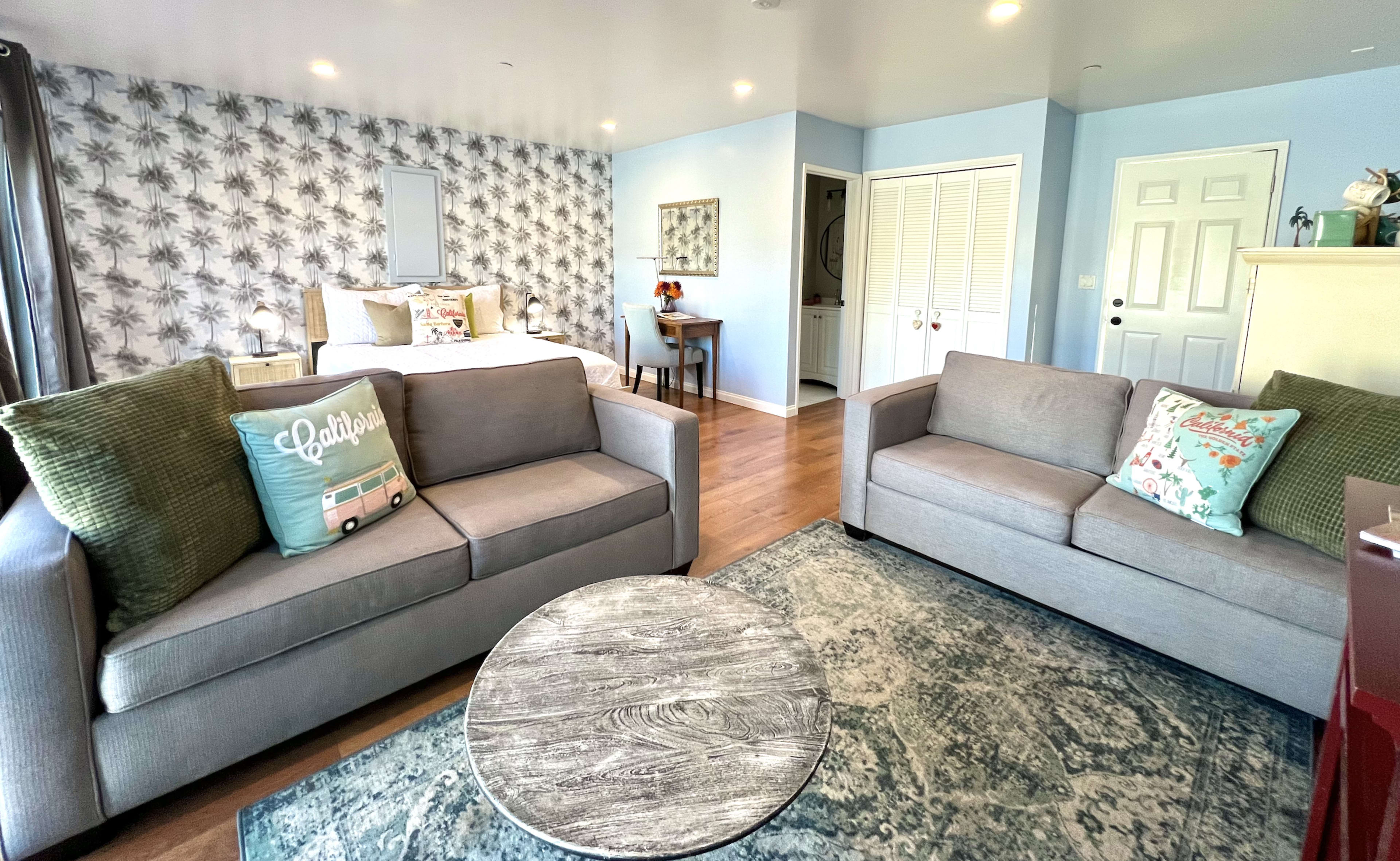 A cozy living space with two gray sofas, a round coffee table, and a bed in the background, all set against a patterned wall and light blue accents.