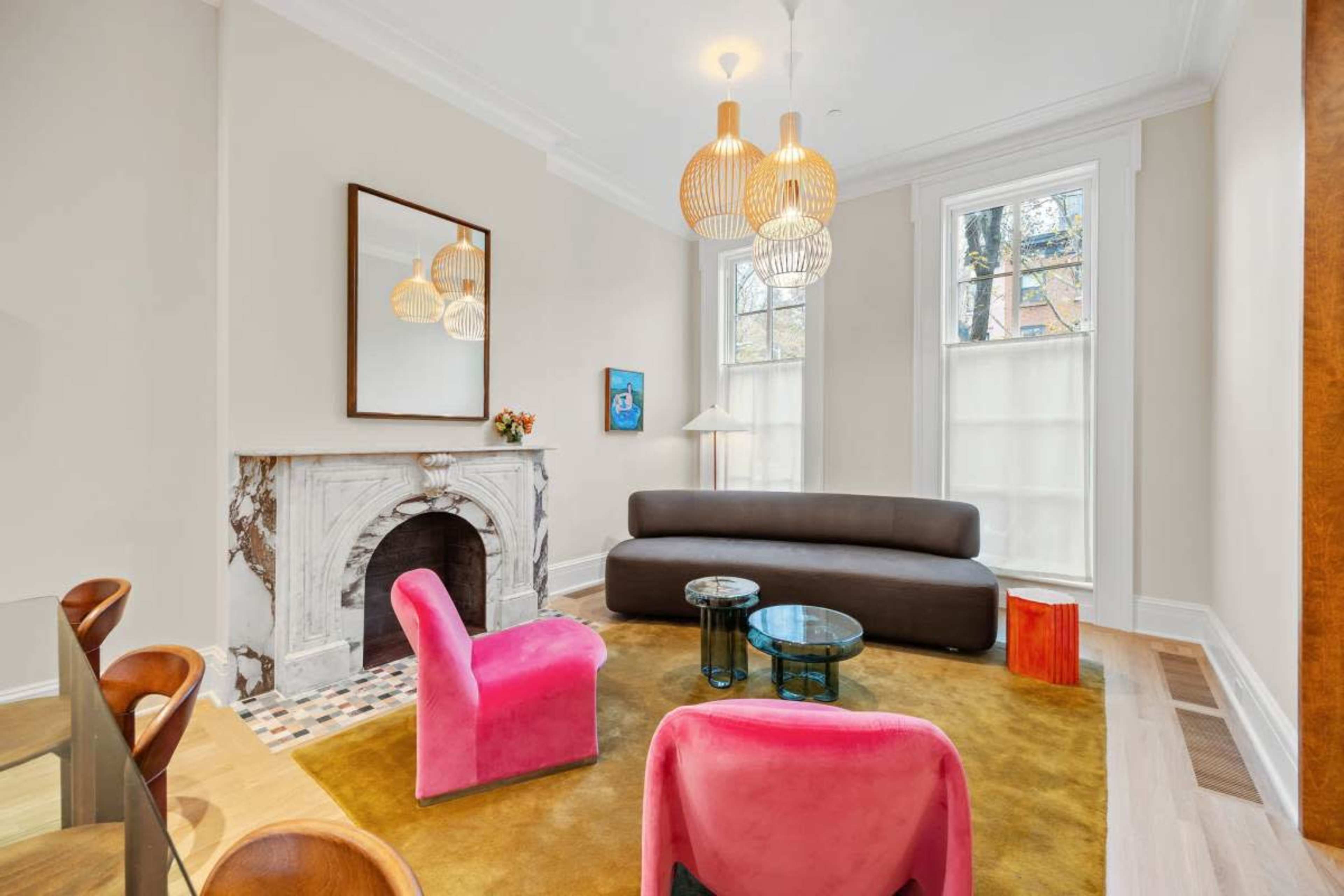 The room features a modern seating arrangement with a brown sofa, pink chairs, and a marble fireplace, illuminated by decorative pendant lights.
