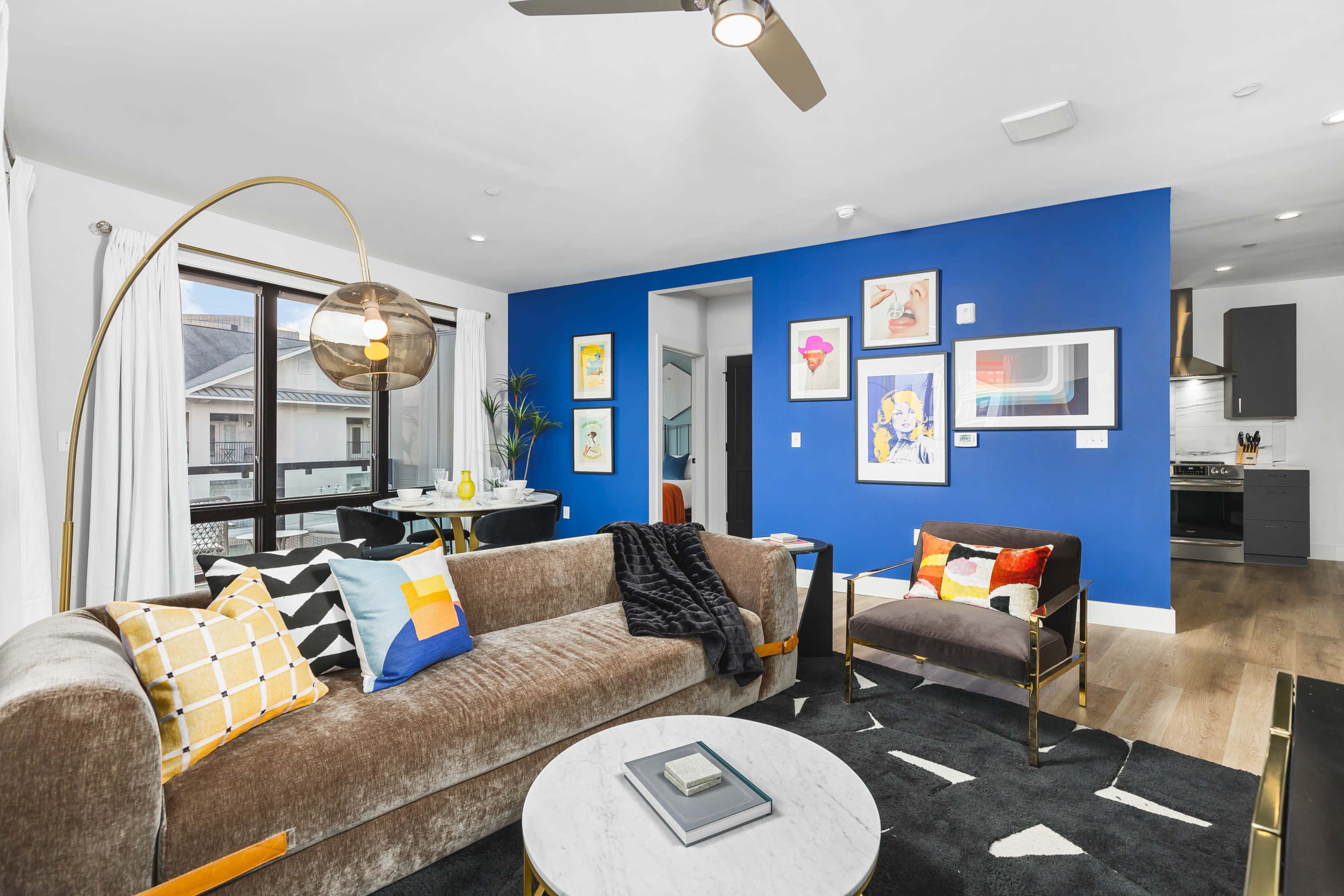 A modern living room featuring a velvet sofa, a round marble coffee table, colorful artwork on blue walls, and a ceiling fan.