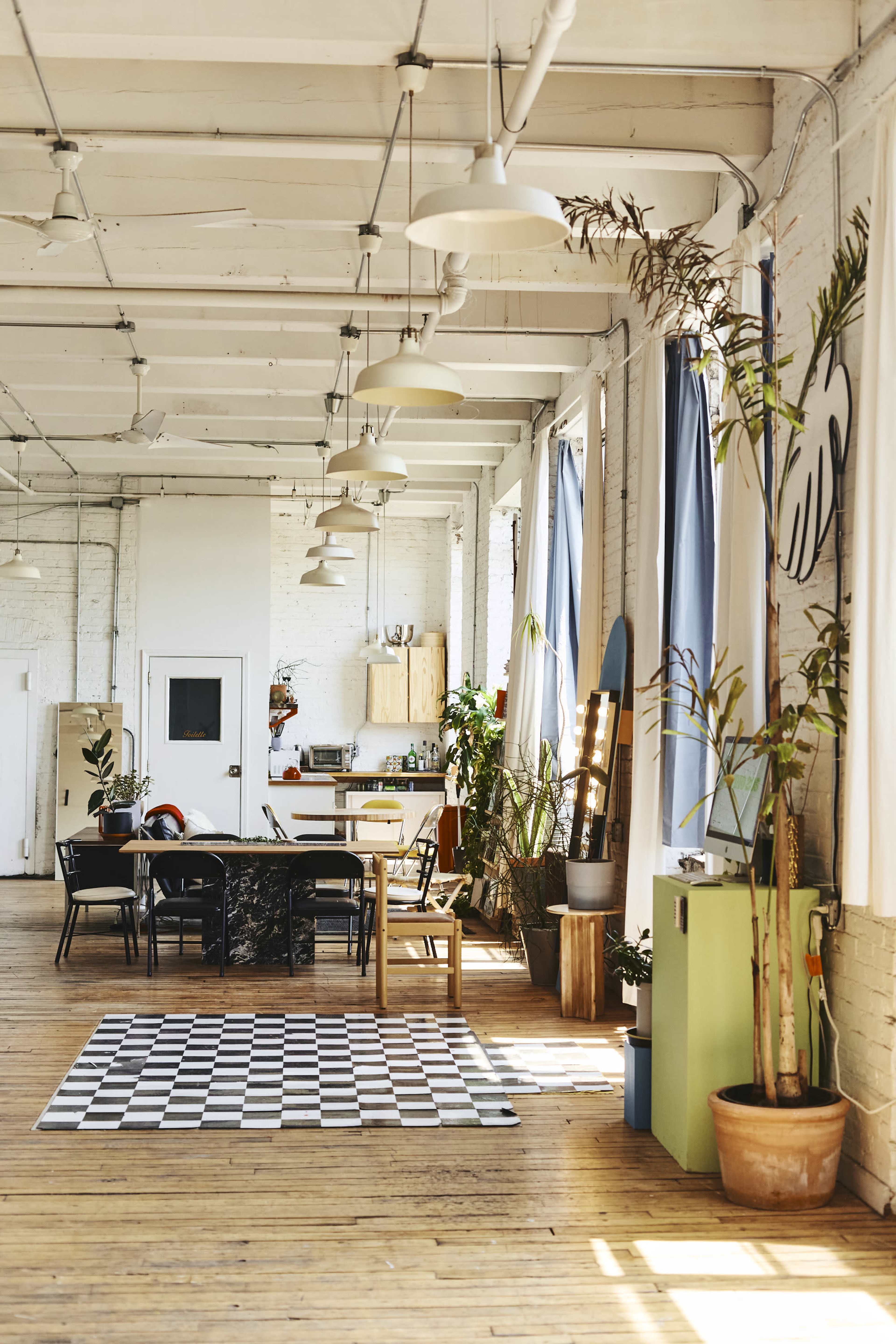 The image shows a spacious, industrial-style room with wooden floors, a dining area featuring a black-and-white checkered rug, plants, and a kitchen in the background.