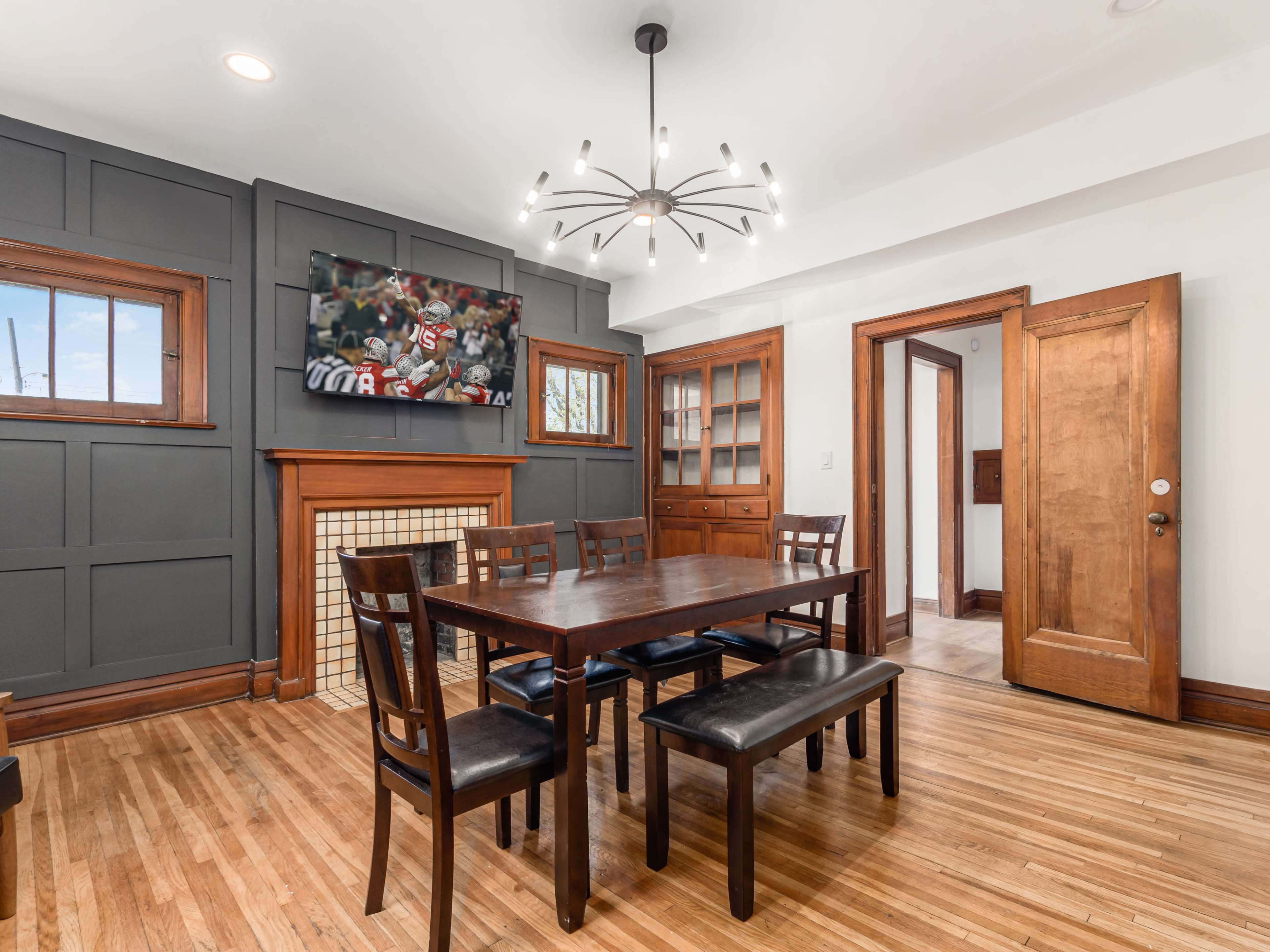 A dining area features a wooden table surrounded by chairs, with a television mounted on the wall and a doorway leading to another room.