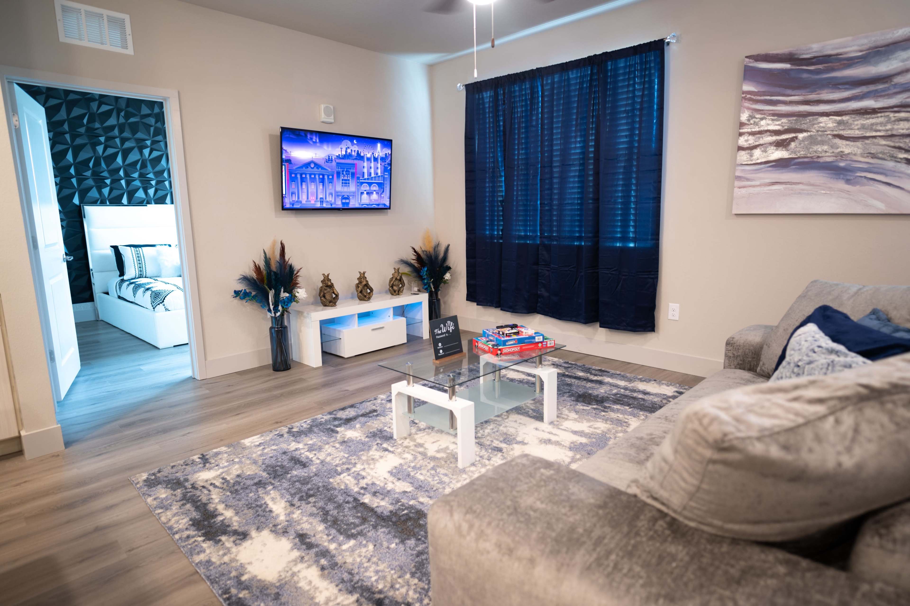 The image shows a modern, brightly lit living room featuring a gray sofa, a glass coffee table, and a large television mounted on the wall, with a blue and white color scheme visible in the decor.