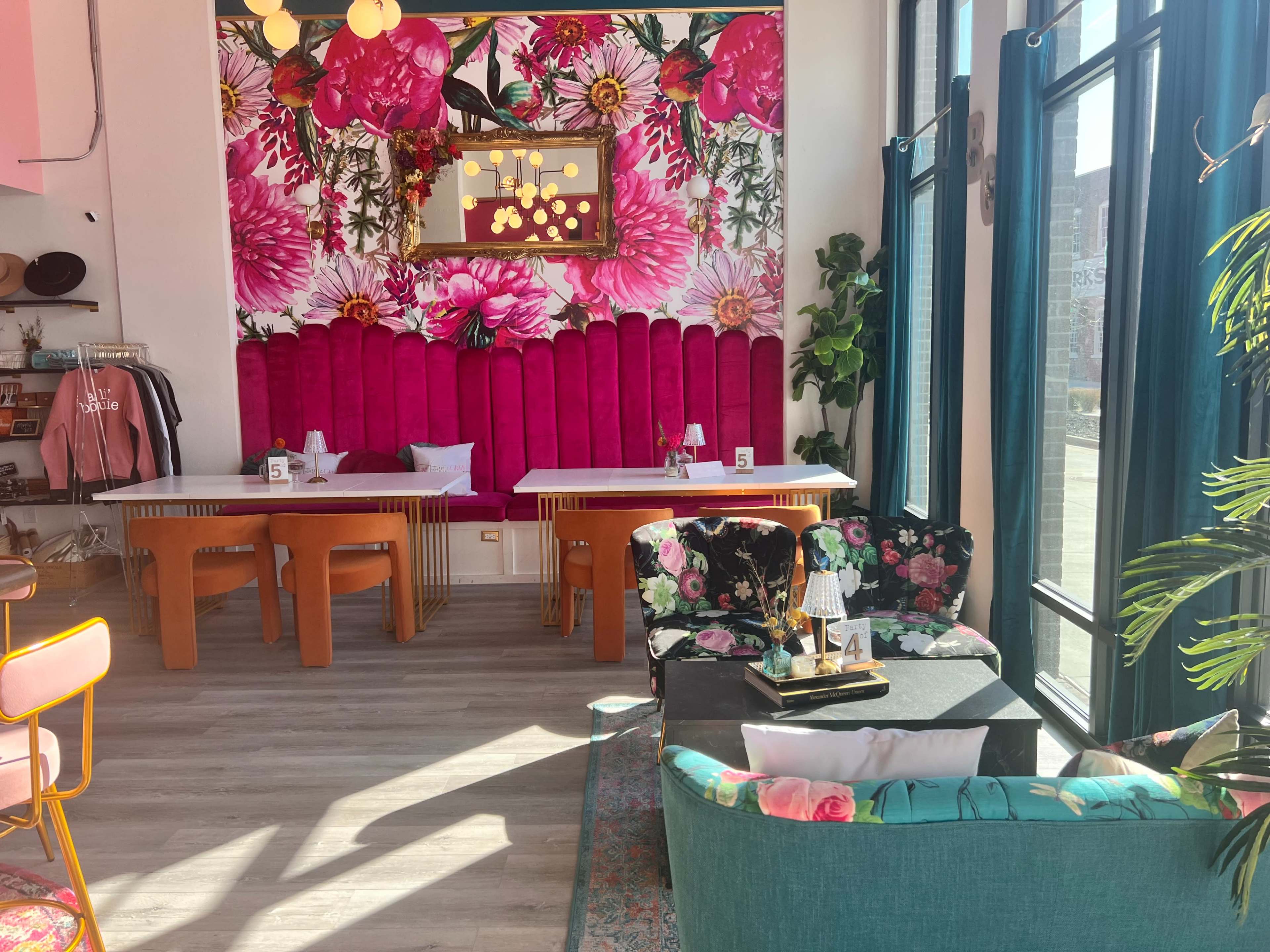 A vibrant café interior with floral wallpaper, two large communal tables, and stylish seating arrangements featuring colorful chairs and cushions.