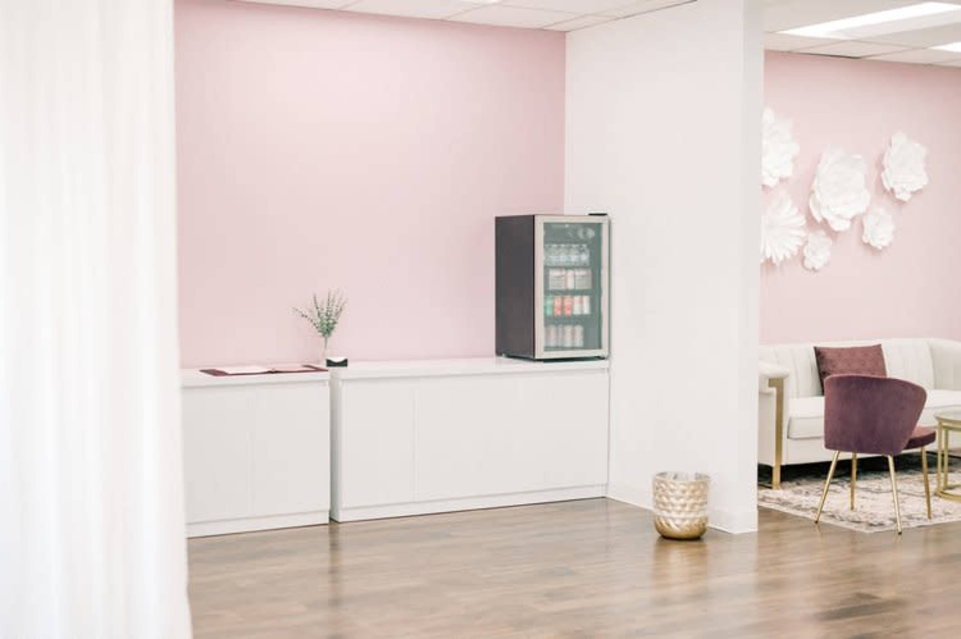 The image shows a modern interior space featuring a pink wall, a drink refrigerator, a white storage cabinet, and a seating area with a cream sofa and a purple chair.