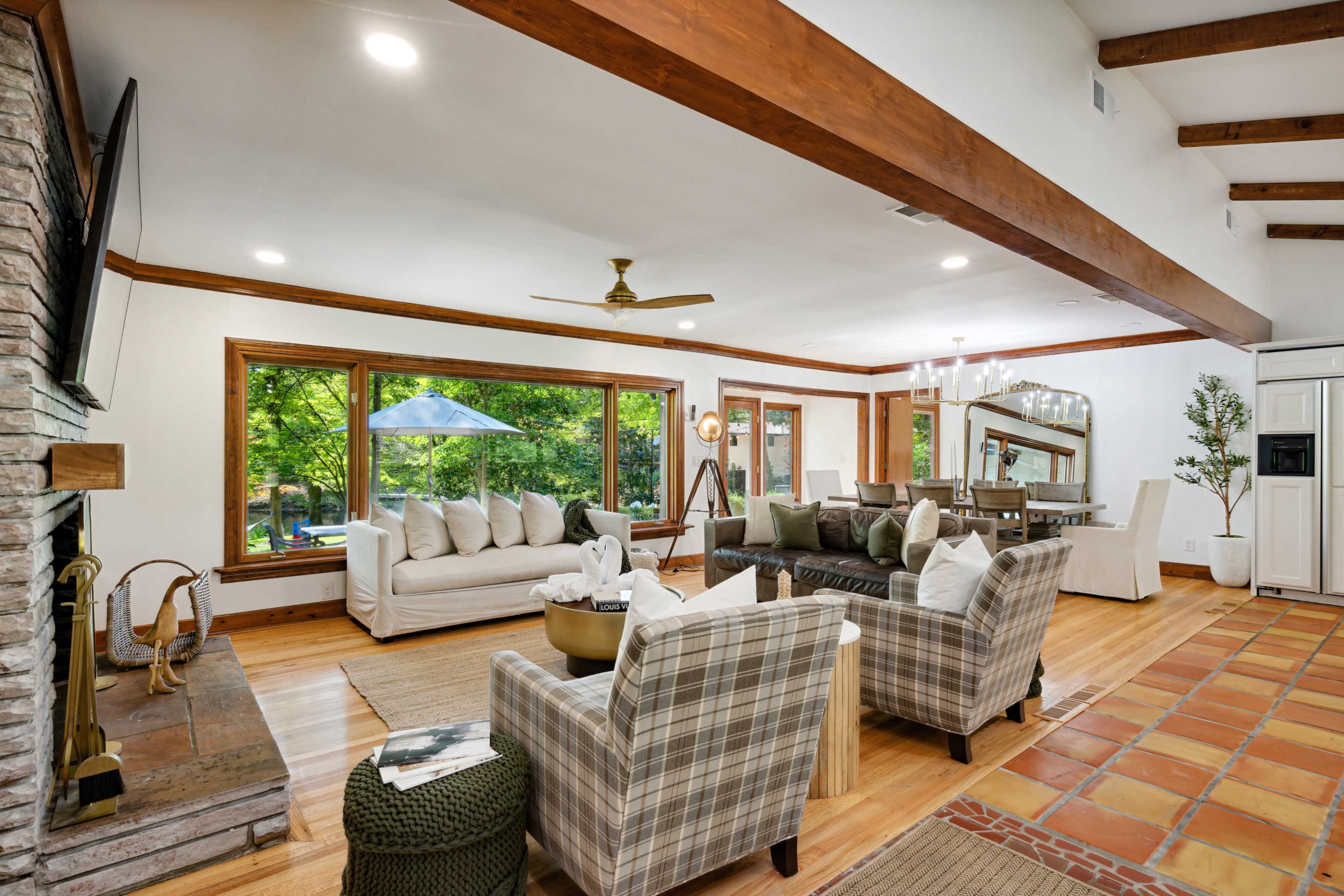 The image shows a spacious living room with large windows overlooking a green outdoor area, featuring a mix of seating arrangements, a stone fireplace, and hardwood floors.
