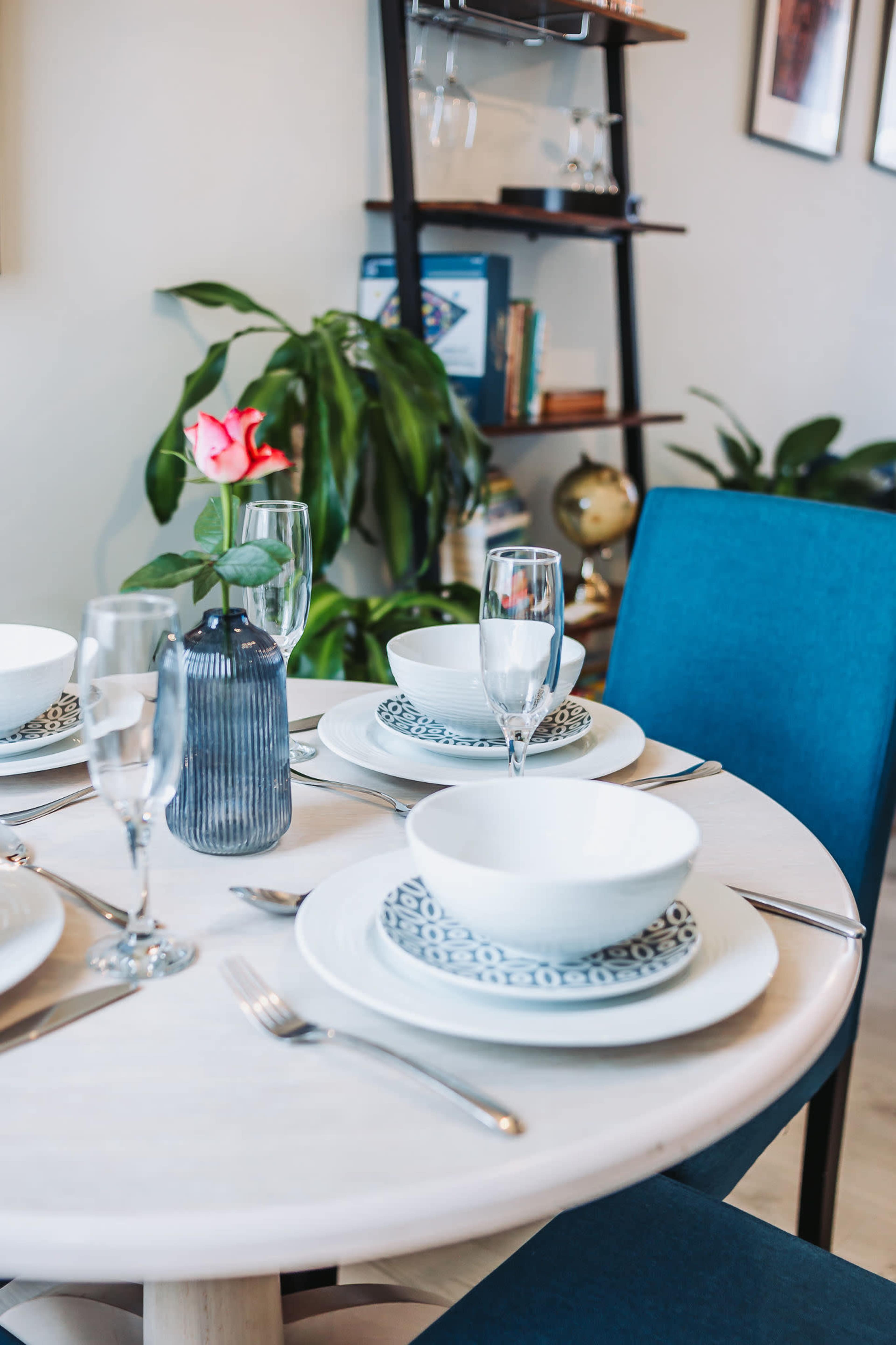 A round dining table is set with white bowls, decorative plates, and glassware, accompanied by a vibrant rose in a vase, against a backdrop of a bookshelf and indoor plants.