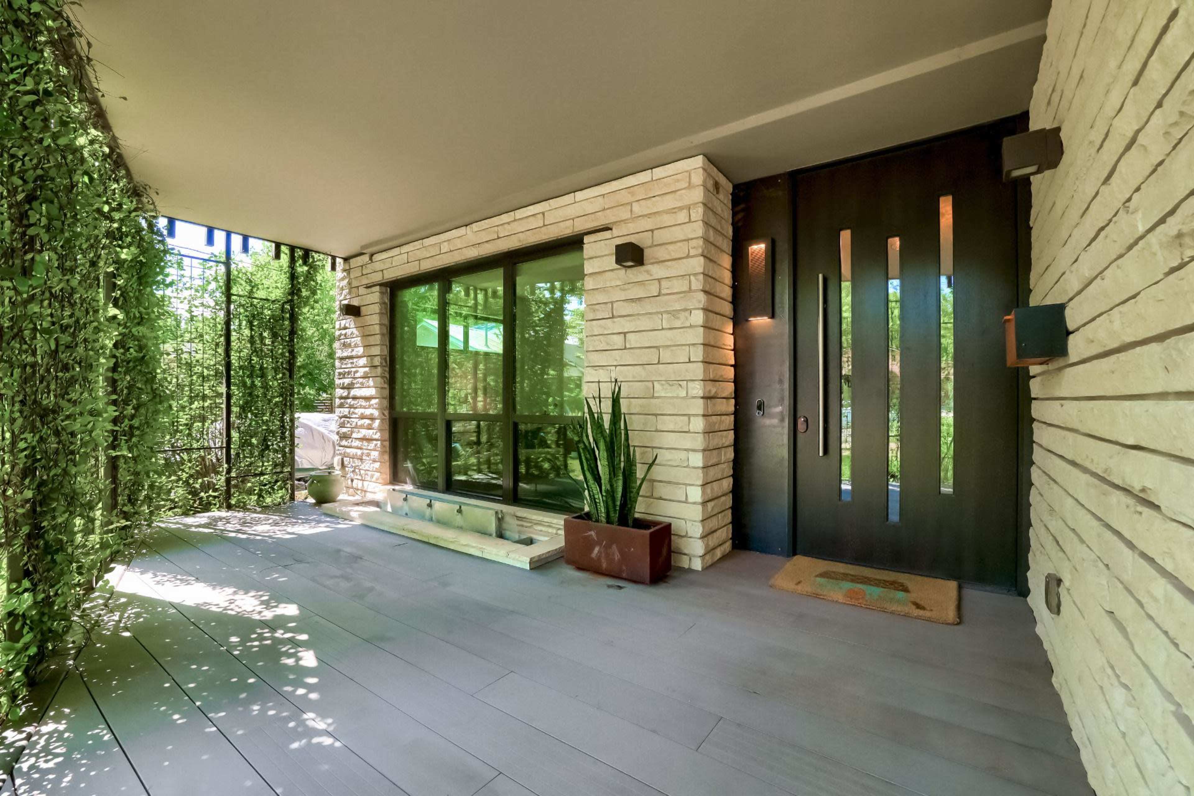A modern entrance with a large wooden door, flanked by glass panels, and surrounded by greenery and potted plants on a wooden deck.