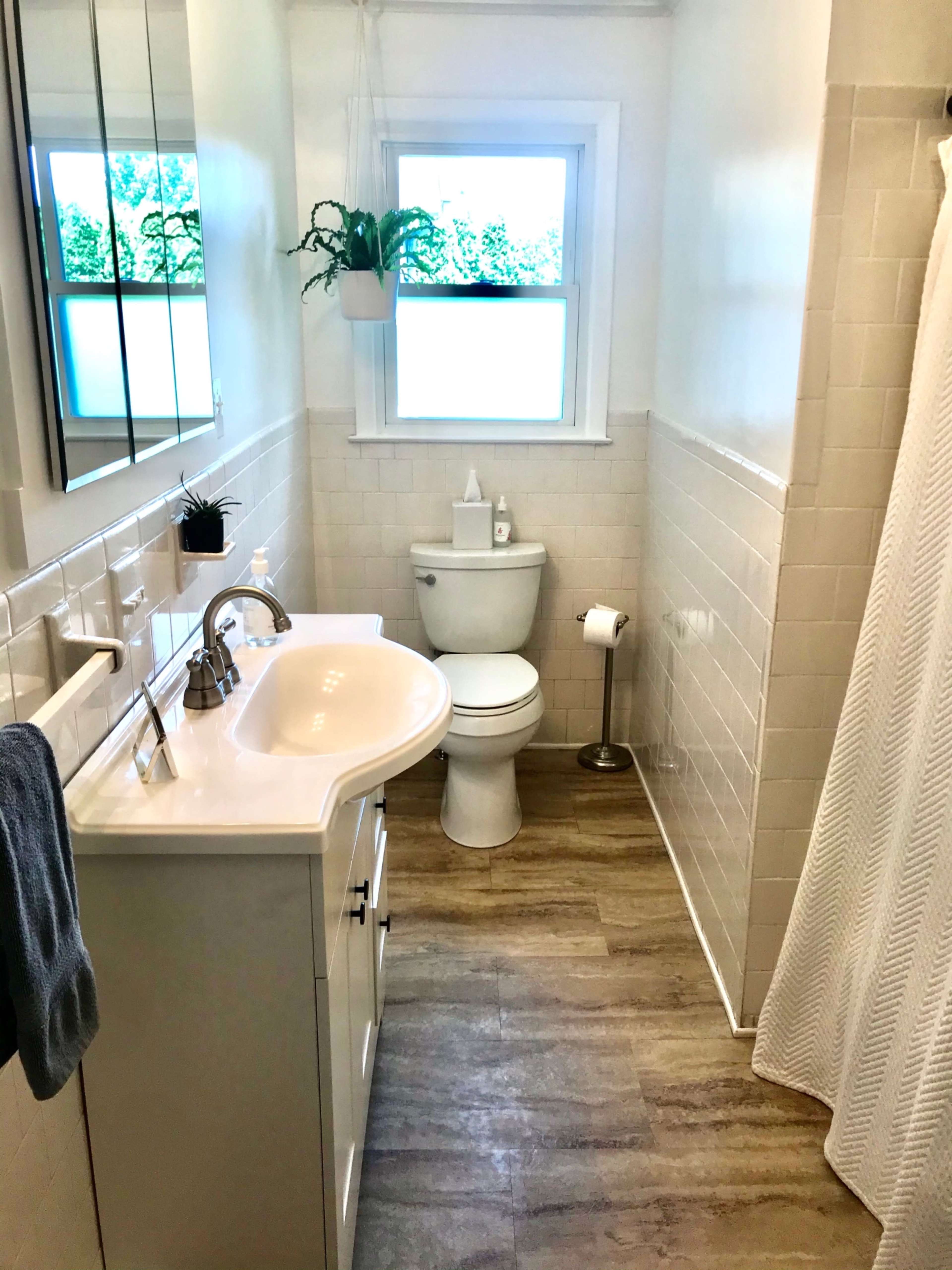 A bright bathroom featuring a white sink with a mirror, a toilet, and a shower curtain, all set against tiled walls and a window allowing natural light.