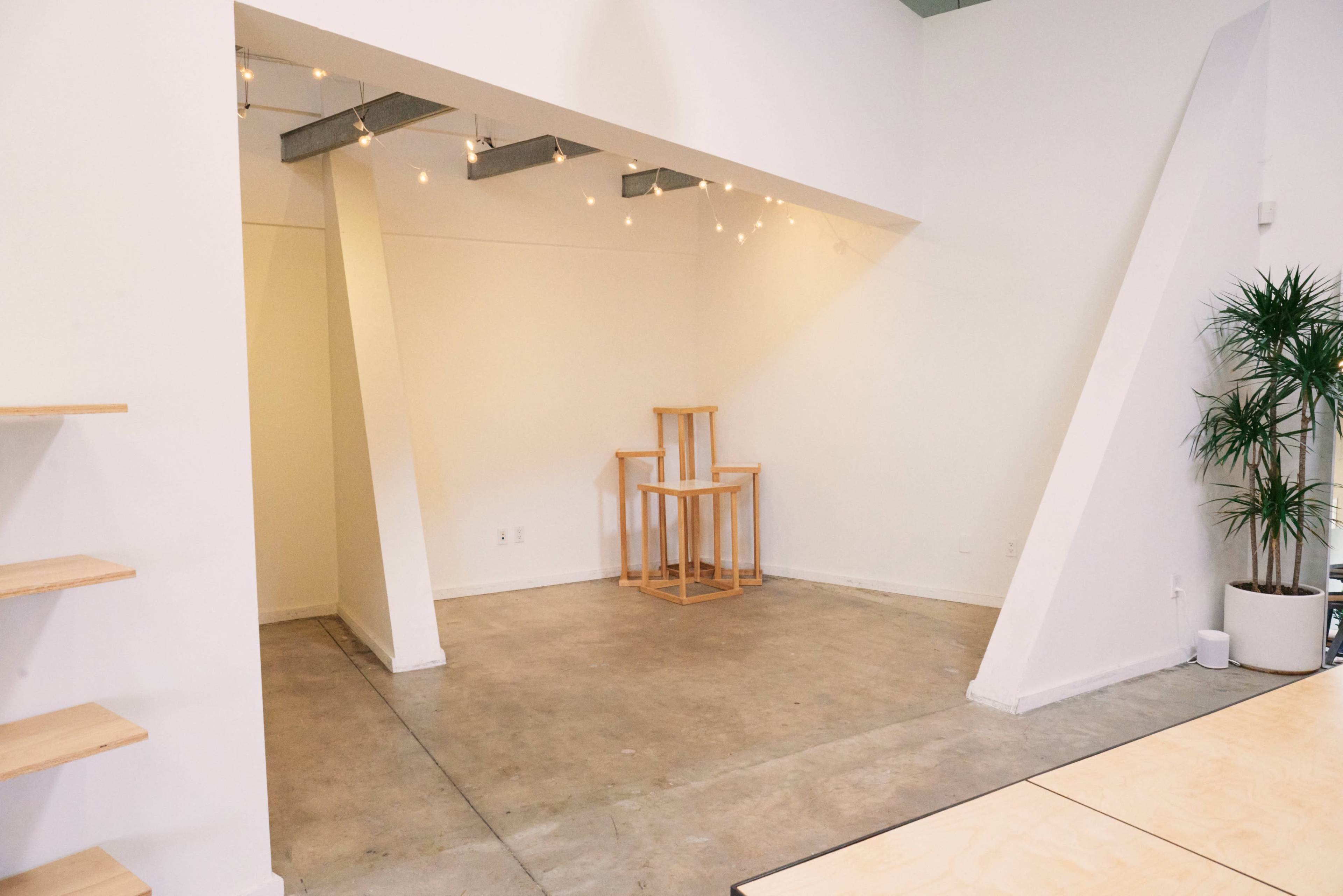 The image shows a minimalist interior space with a concrete floor, white walls, and a few wooden furniture pieces arranged in the center, illuminated by string lights overhead.