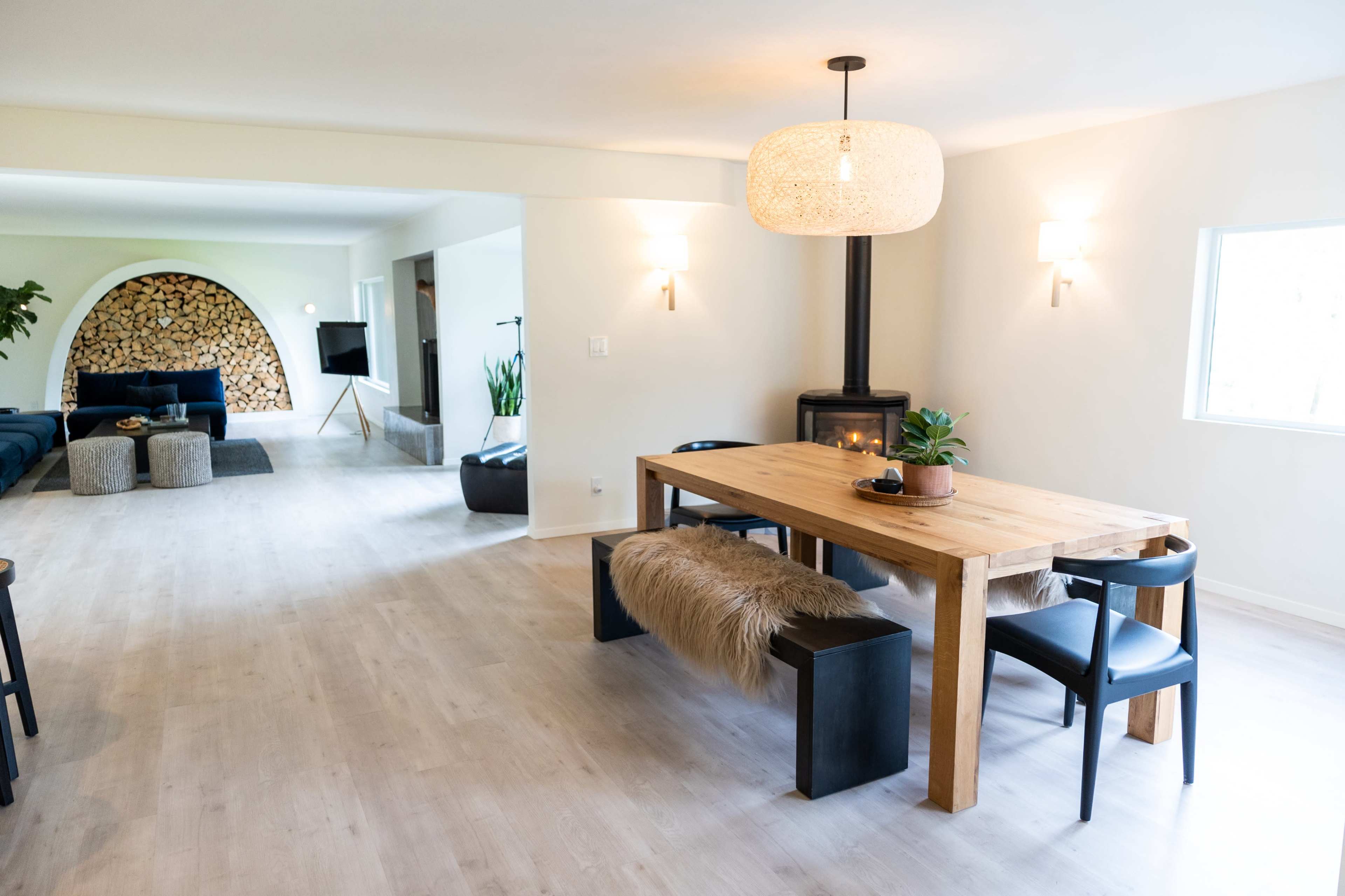The image shows a modern dining area featuring a wooden table with black chairs and a plant, adjacent to a living space with a large sofa and a decorative wood stack.