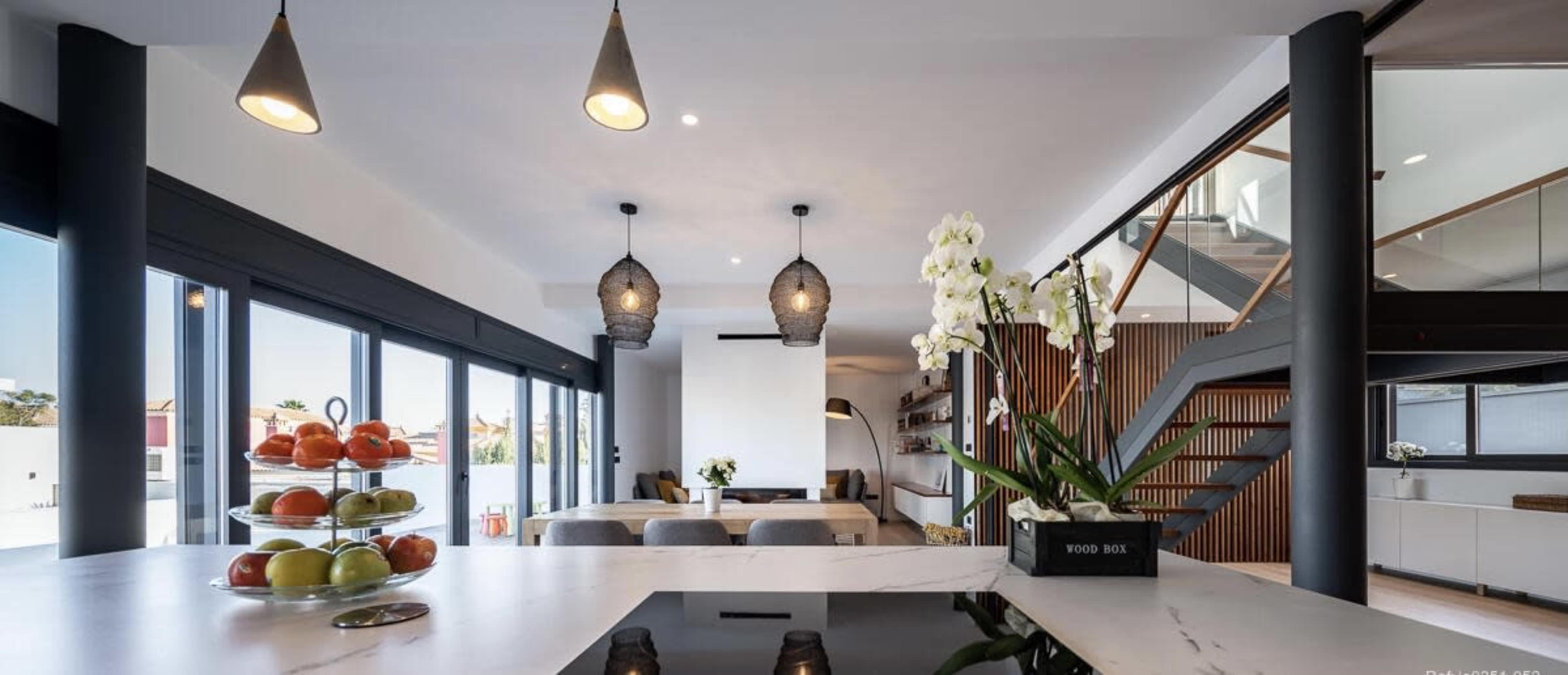 The image shows a modern kitchen and living area, featuring a marble countertop, fruits displayed in a bowl, and a staircase leading to an upper level.