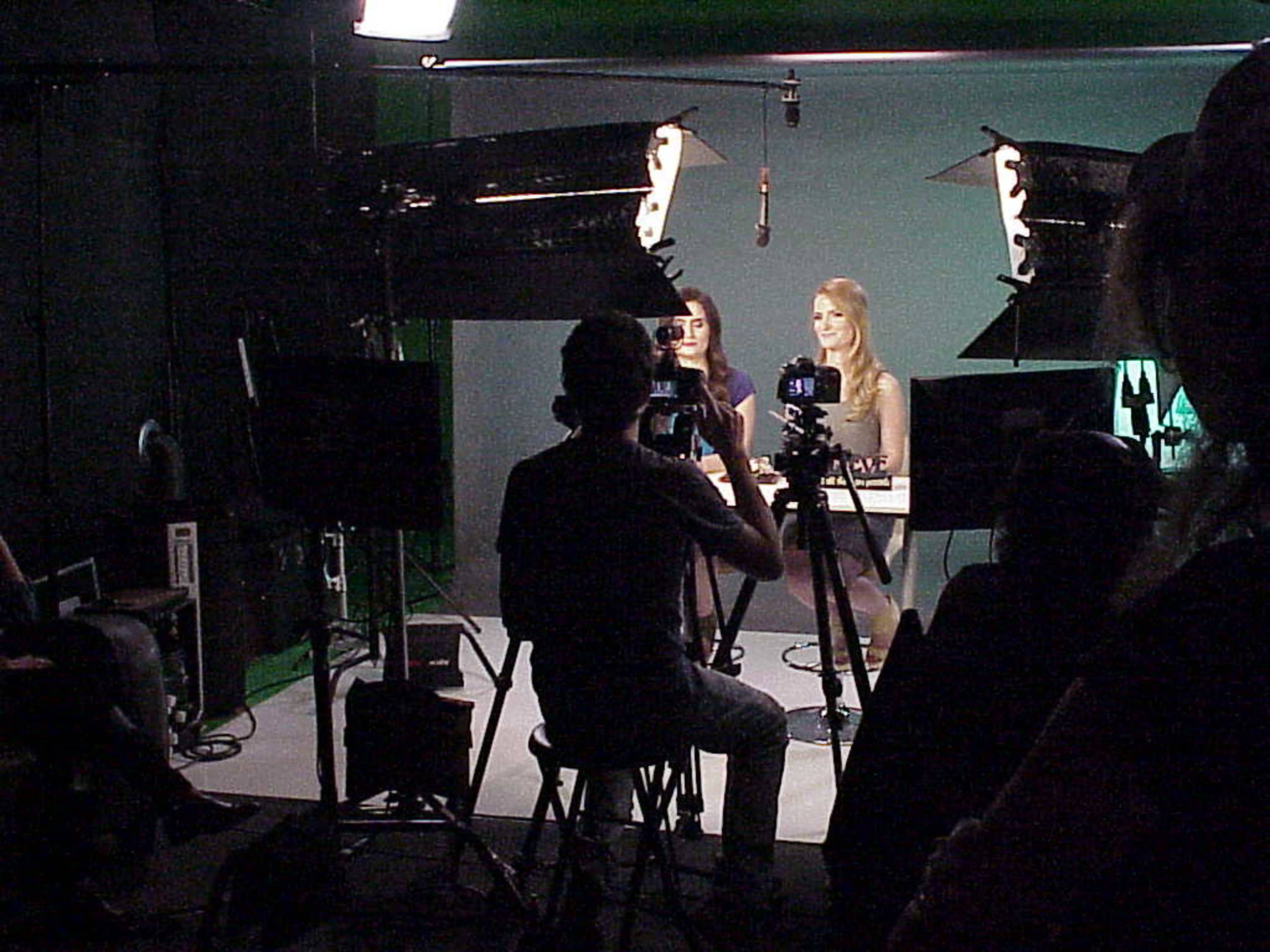 Two women are seated at a table with microphones in front of them, while a person behind a camera captures the scene in a studio setting.