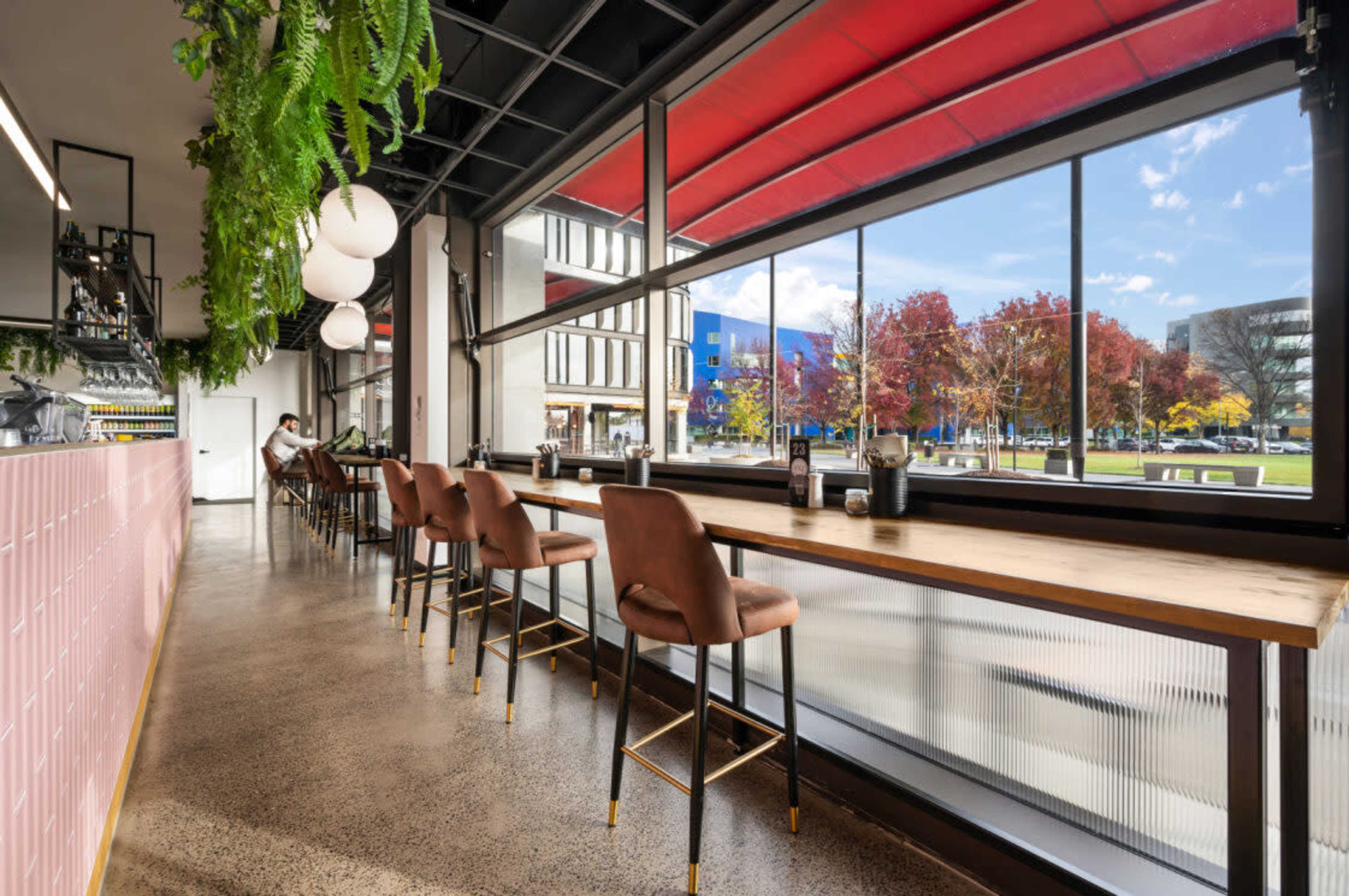 A modern café features a long wooden bar with high stools, large windows overlooking a colorful outdoor scene filled with trees and buildings.