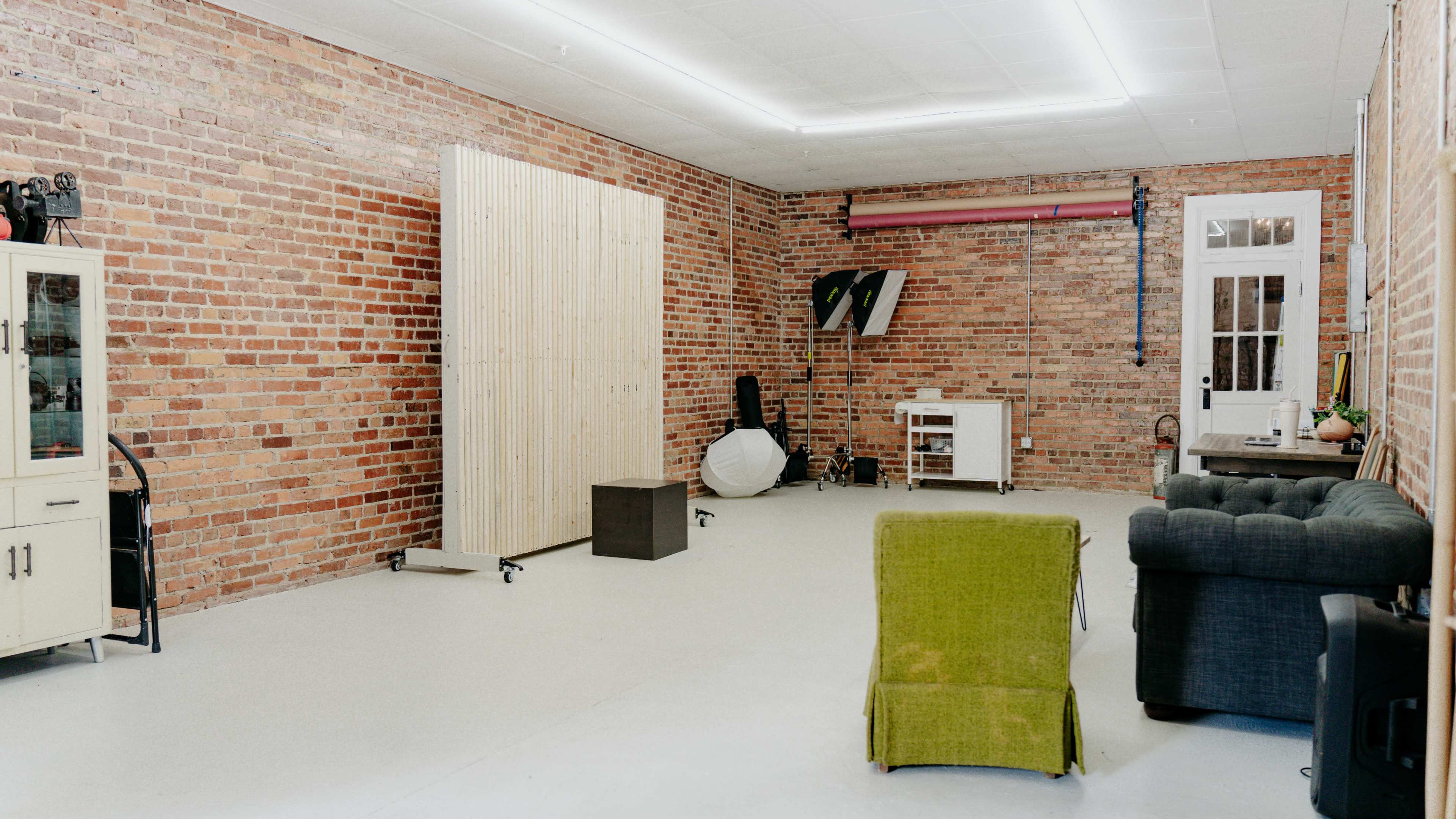 The image shows an empty studio space with exposed brick walls, a wooden partition, and furniture including a green chair and a black couch.