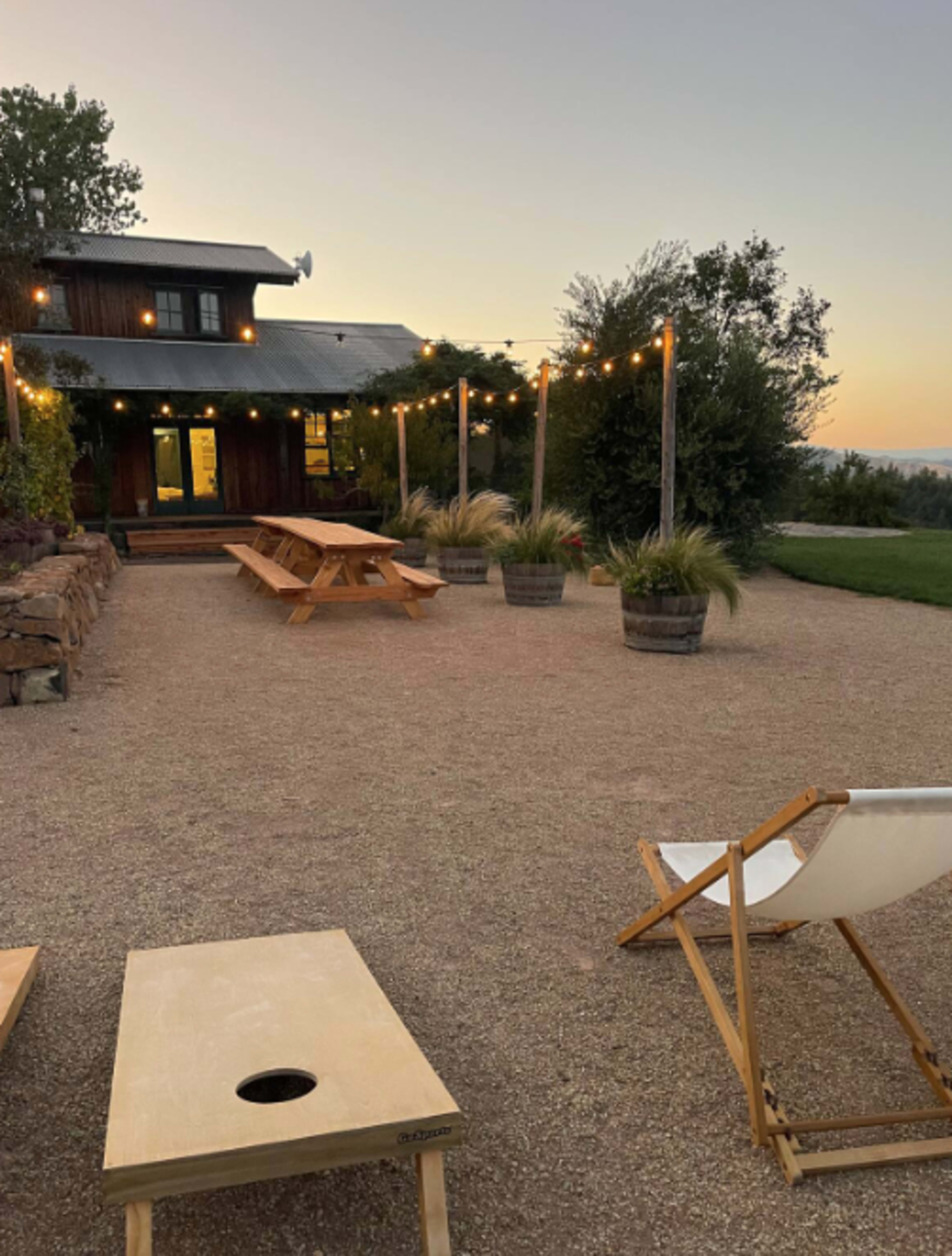 The image shows a gravel outdoor area featuring a wooden picnic table, potted plants, and string lights, with a building in the background at sunset.